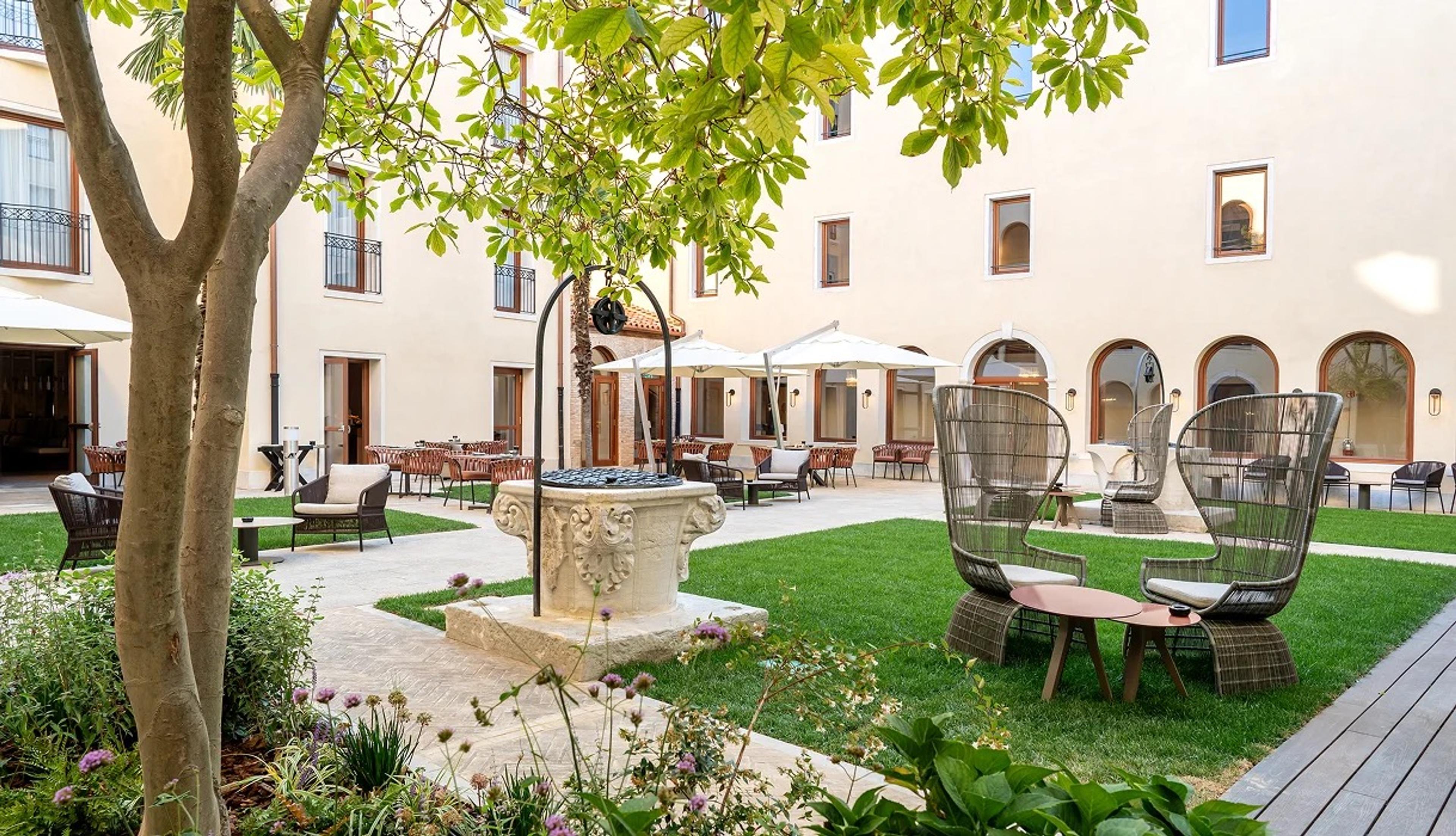 A courtyard garden with a stone wellhead, wicker chairs on the lawn, and arched windows along the building behind.