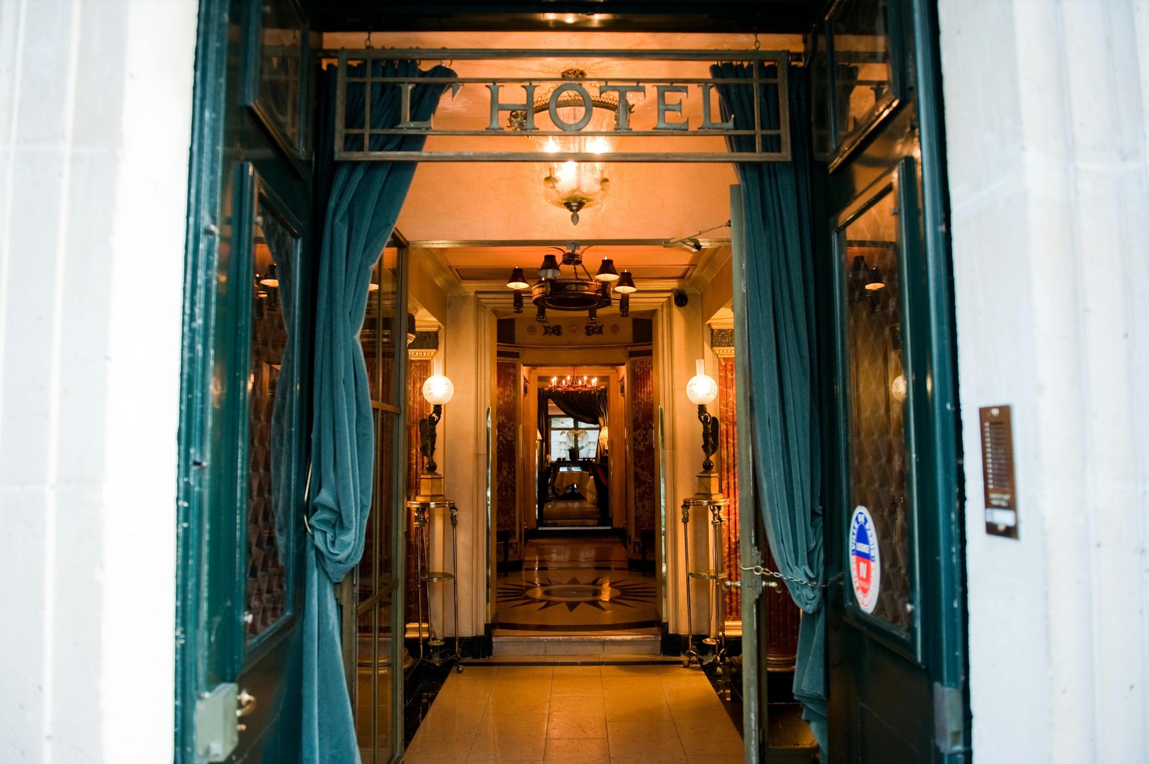 A view down the hotel's teal-curtained entrance corridor, past wall lamps to a sunburst mosaic floor at the far end, with the 'L'Hotel' sign hanging overhead.