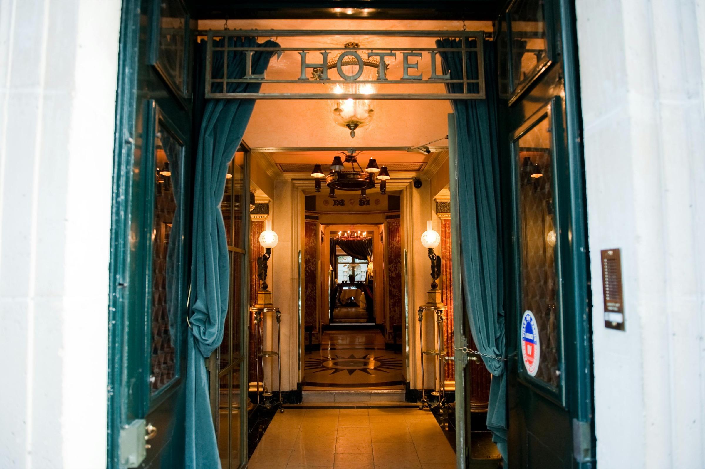 A view down the hotel's teal-curtained entrance corridor, past wall lamps to a sunburst mosaic floor at the far end, with the 'L'Hotel' sign hanging overhead.