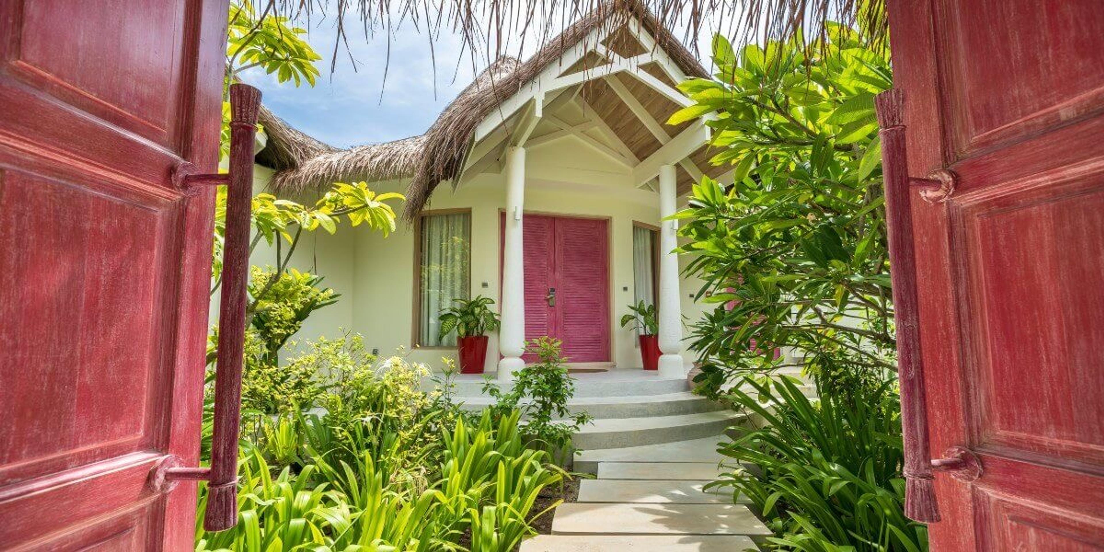 A pair of open red doors framing a view through a tropical garden to a thatched villa entrance.