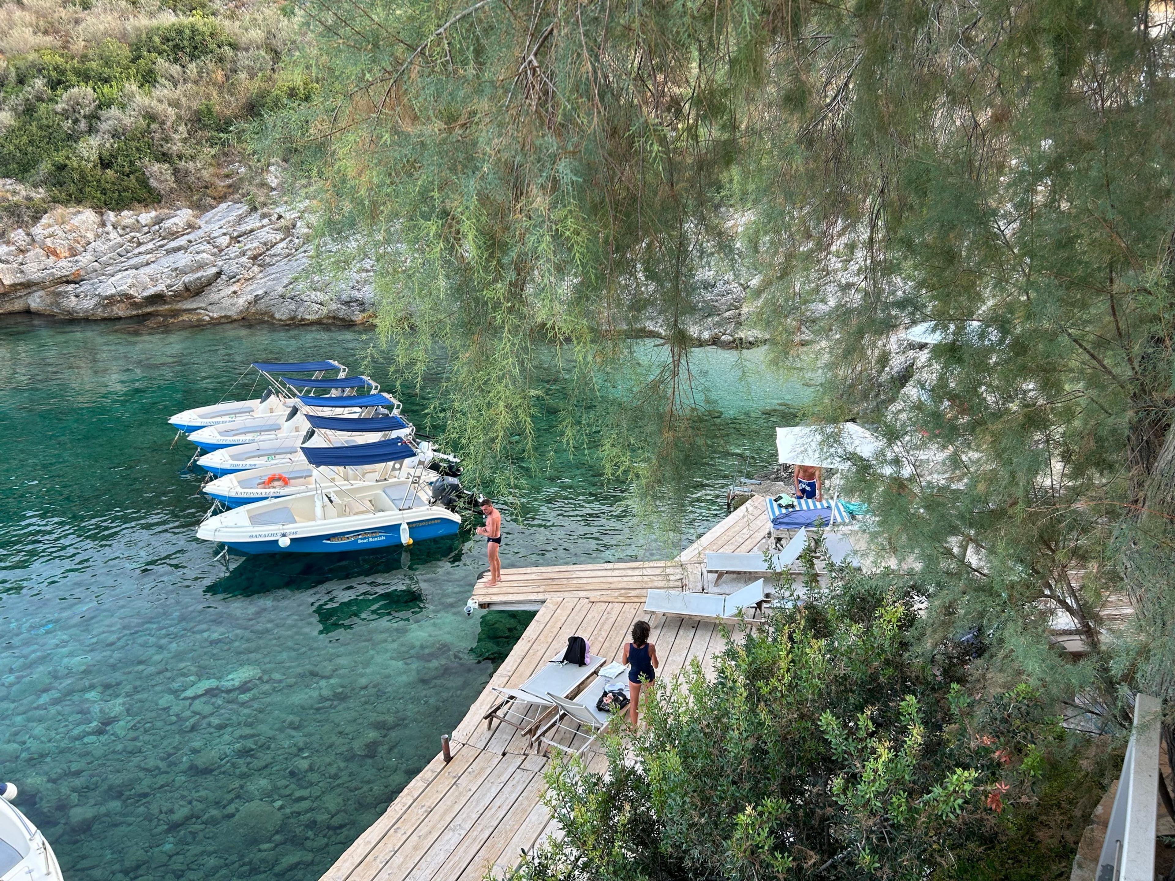 Clear turquoise water over pebbles in a small rocky cove near Agios Nikolaos, with a row of motorboats moored beside a timber jetty and sunloungers, seen through tamarisk branches