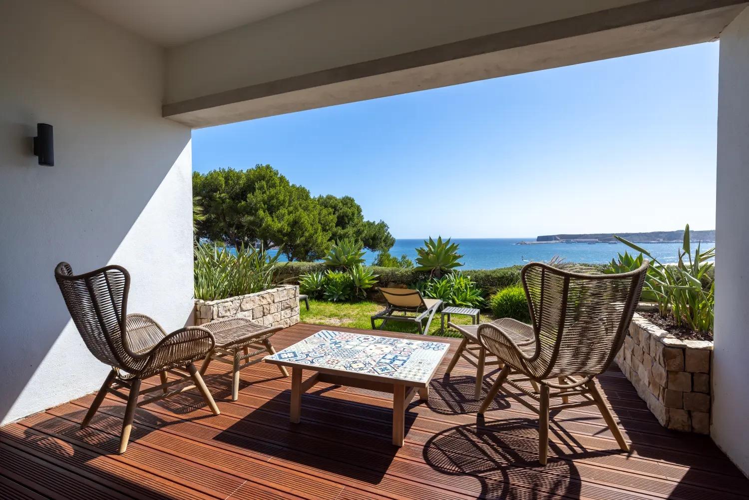 A covered terrace outside a room, with two woven chairs and a tiled table, a lounger on the grass beyond and the sea past the trees.
