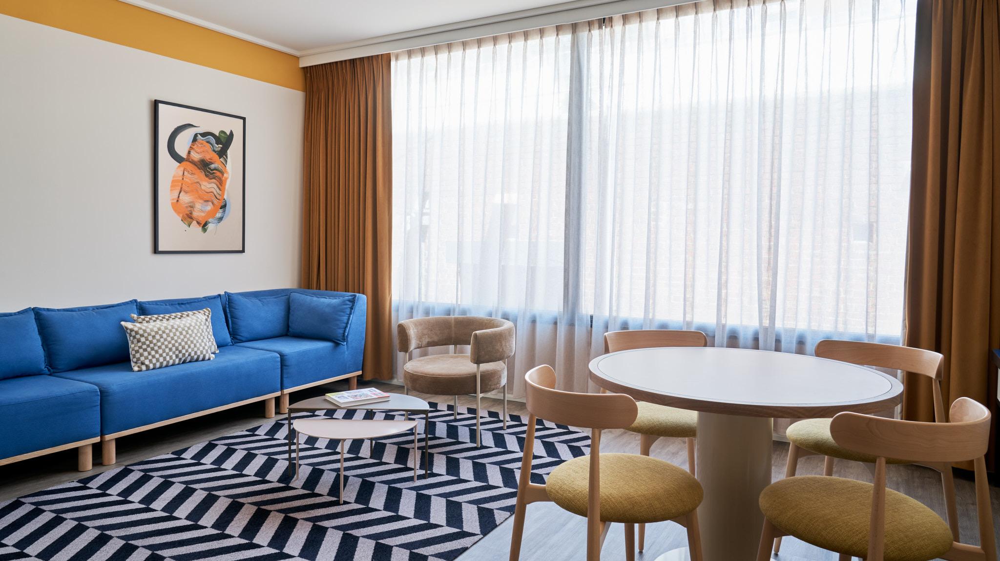 A hotel living room with a blue corner sofa, a round table and chair, a wall-mounted television and floor-to-ceiling curtained windows.