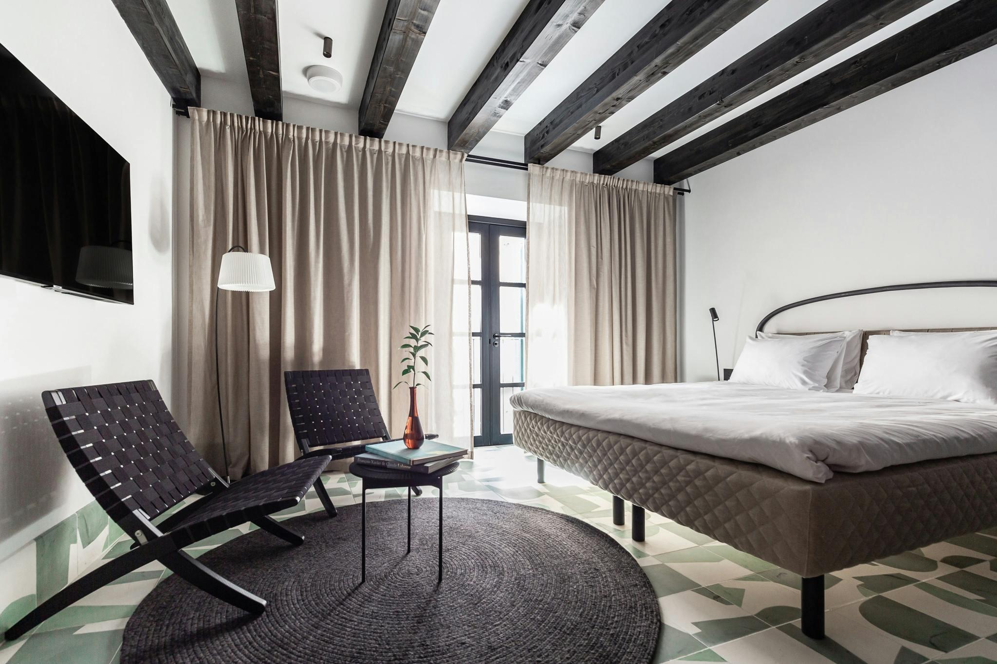 A bedroom with dark ceiling beams, a bed dressed in white linen, two woven lounge chairs around a low table, and a green and white patterned tile floor.
