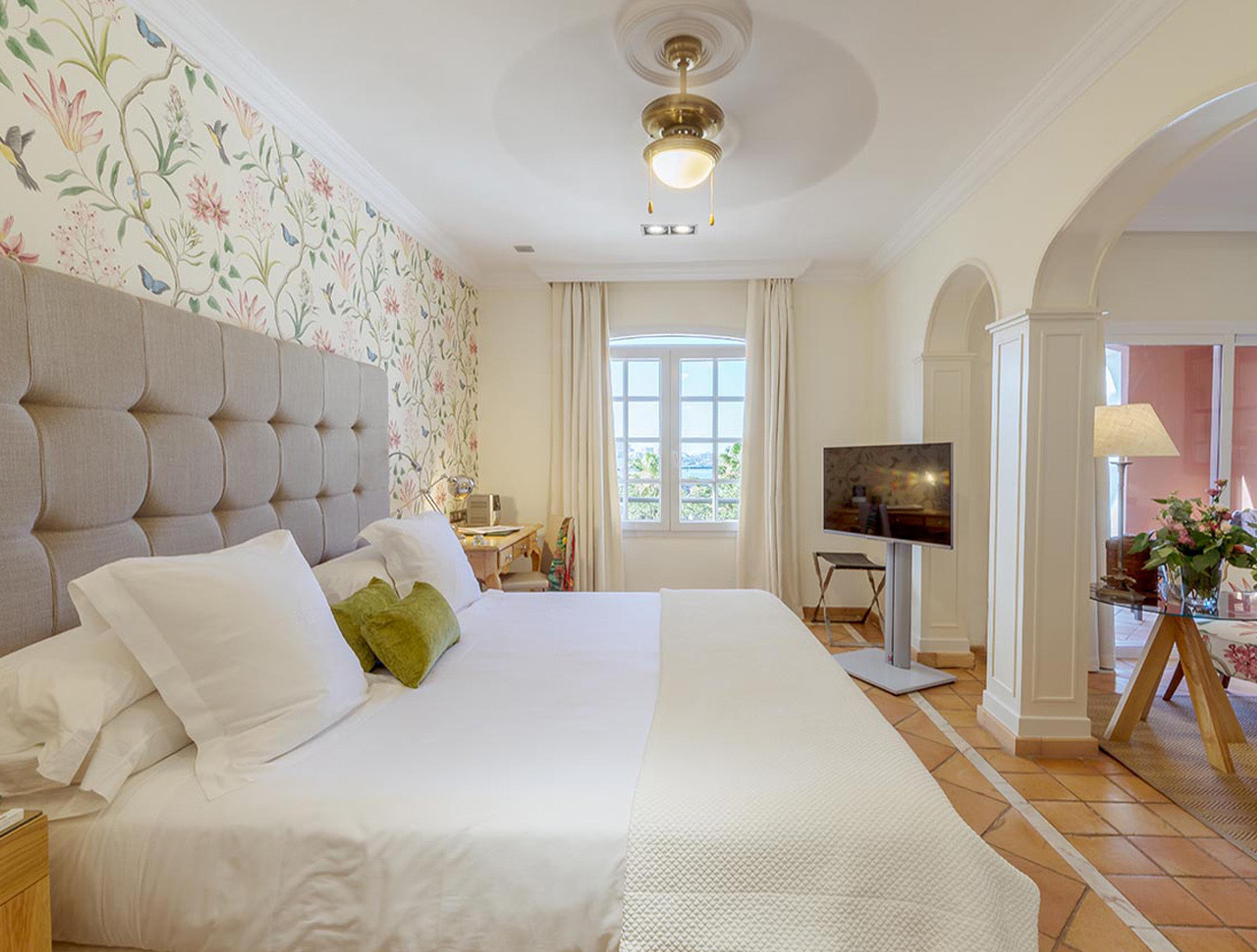 A bedroom with floral wallpaper behind a grey upholstered headboard, fresh flowers on a side table and an archway opening into a sitting room.