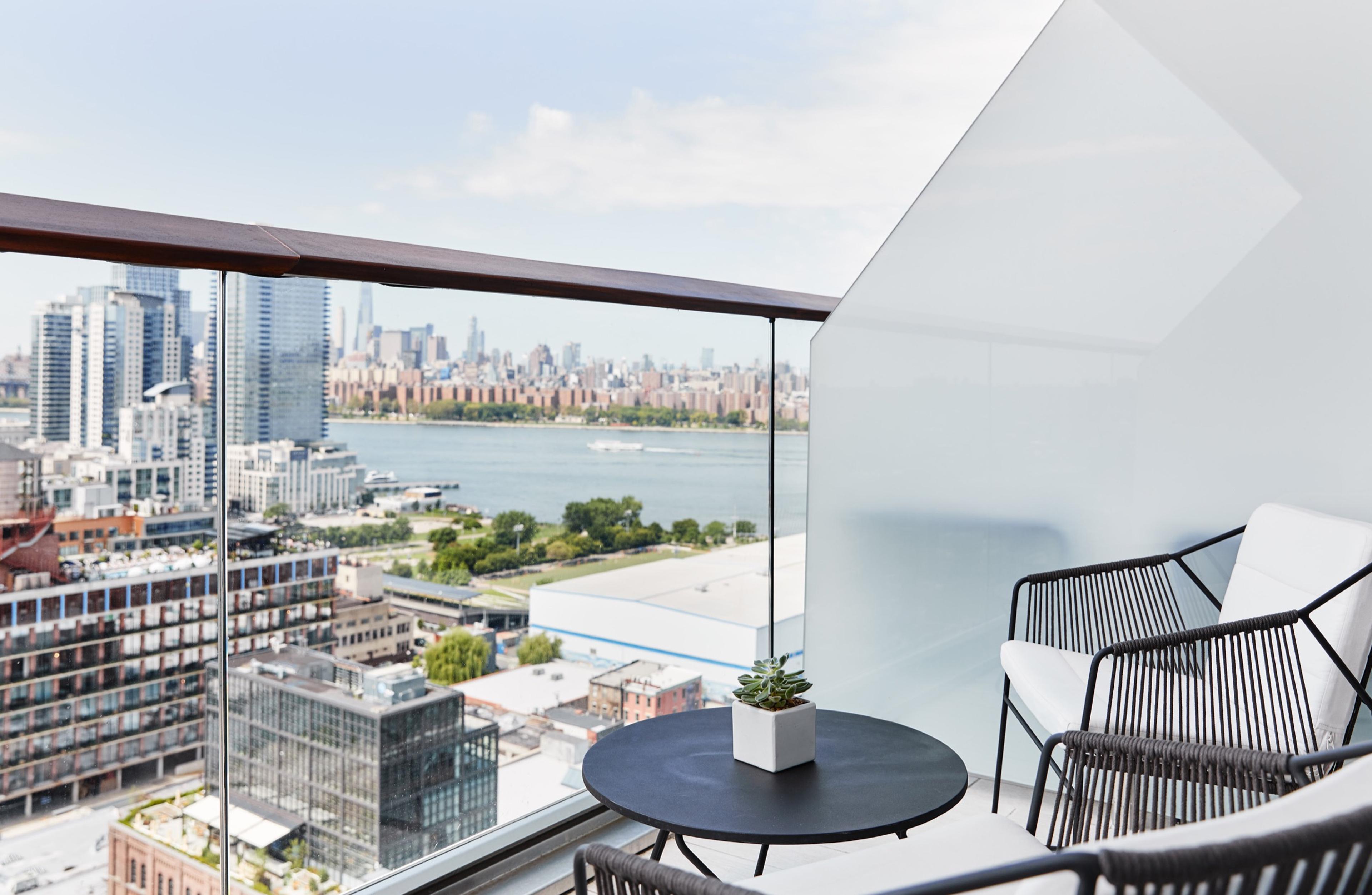 A balcony with woven chairs around a small round table, overlooking a river, apartment towers, and the Manhattan skyline.