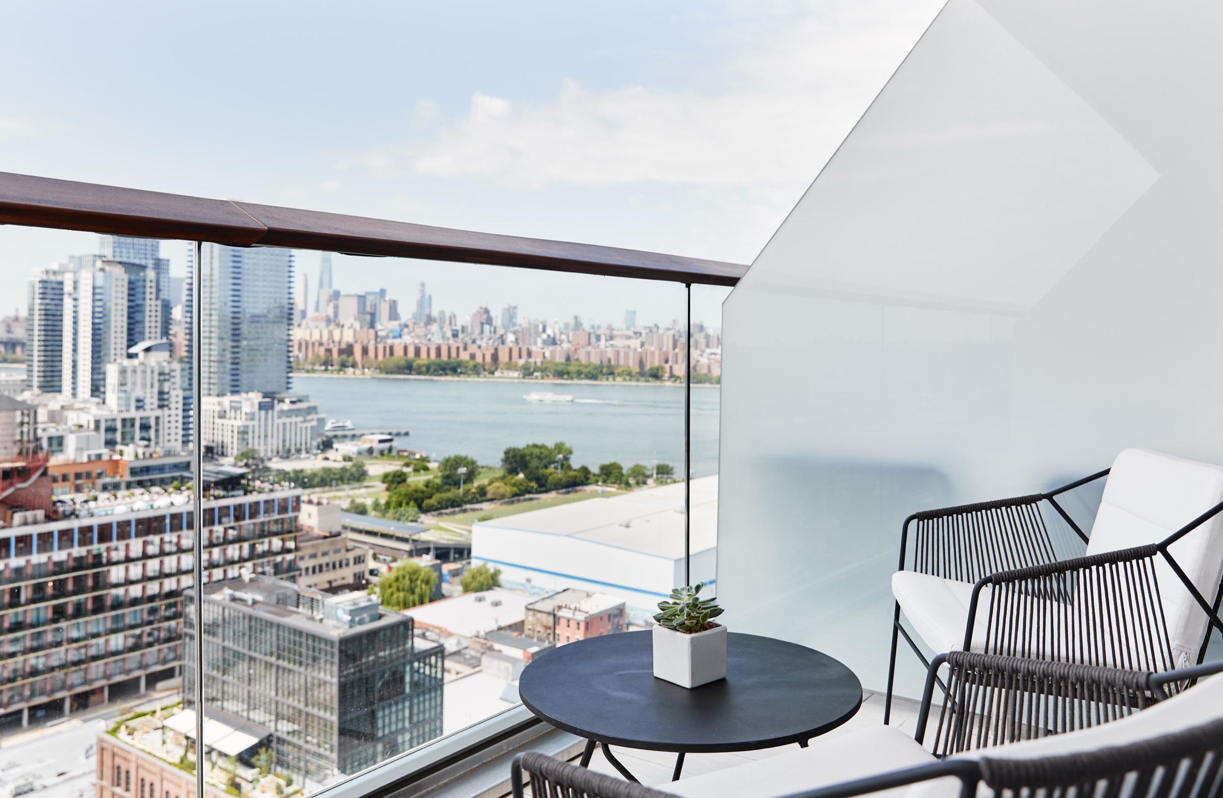 A balcony with woven chairs around a small round table, overlooking a river, apartment towers, and the Manhattan skyline.
