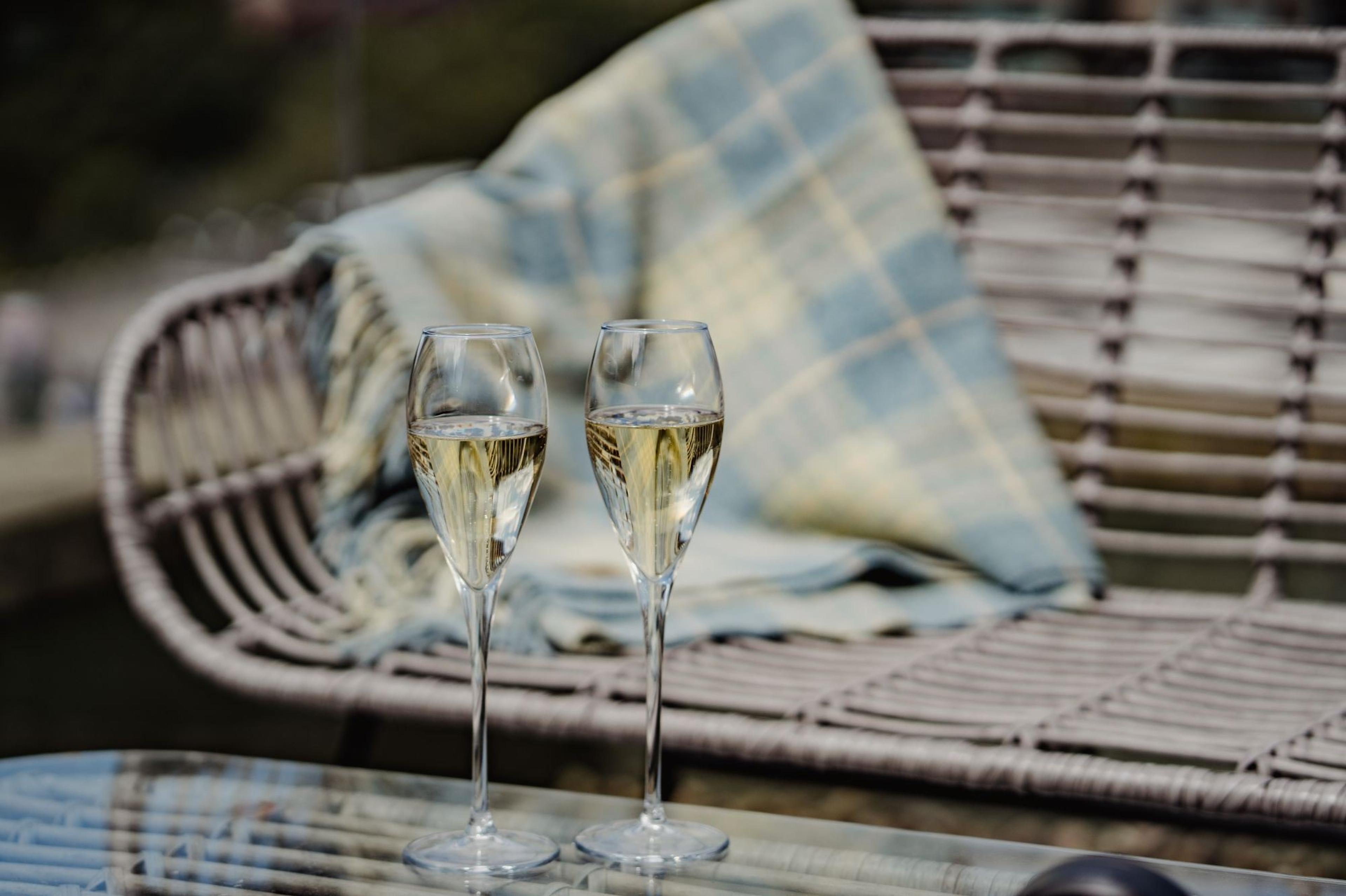 Two glasses of champagne on an outdoor table beside a wicker chair with a wool blanket, the city blurred in the background.