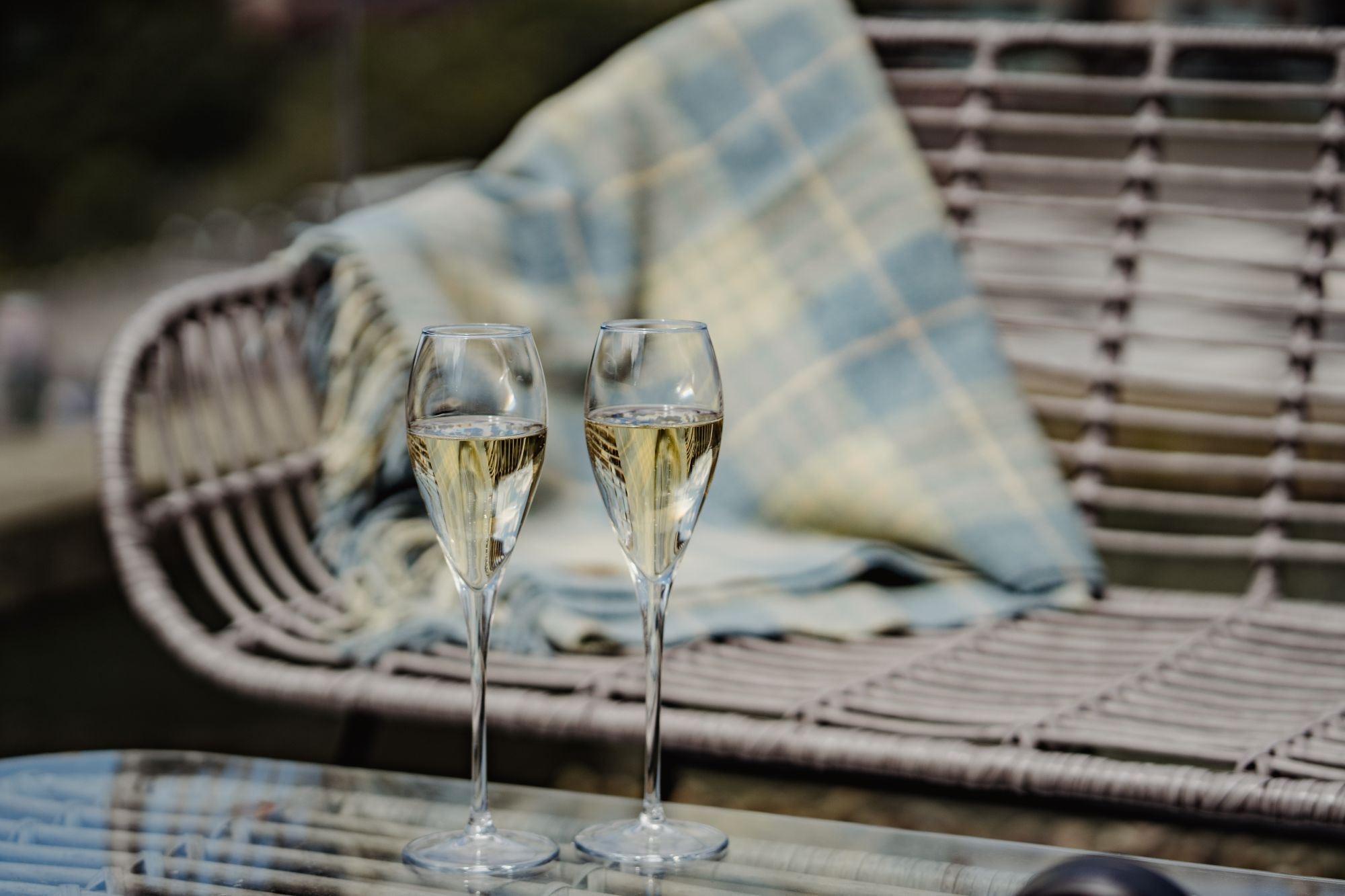 Two glasses of champagne on an outdoor table beside a wicker chair with a wool blanket, the city blurred in the background.