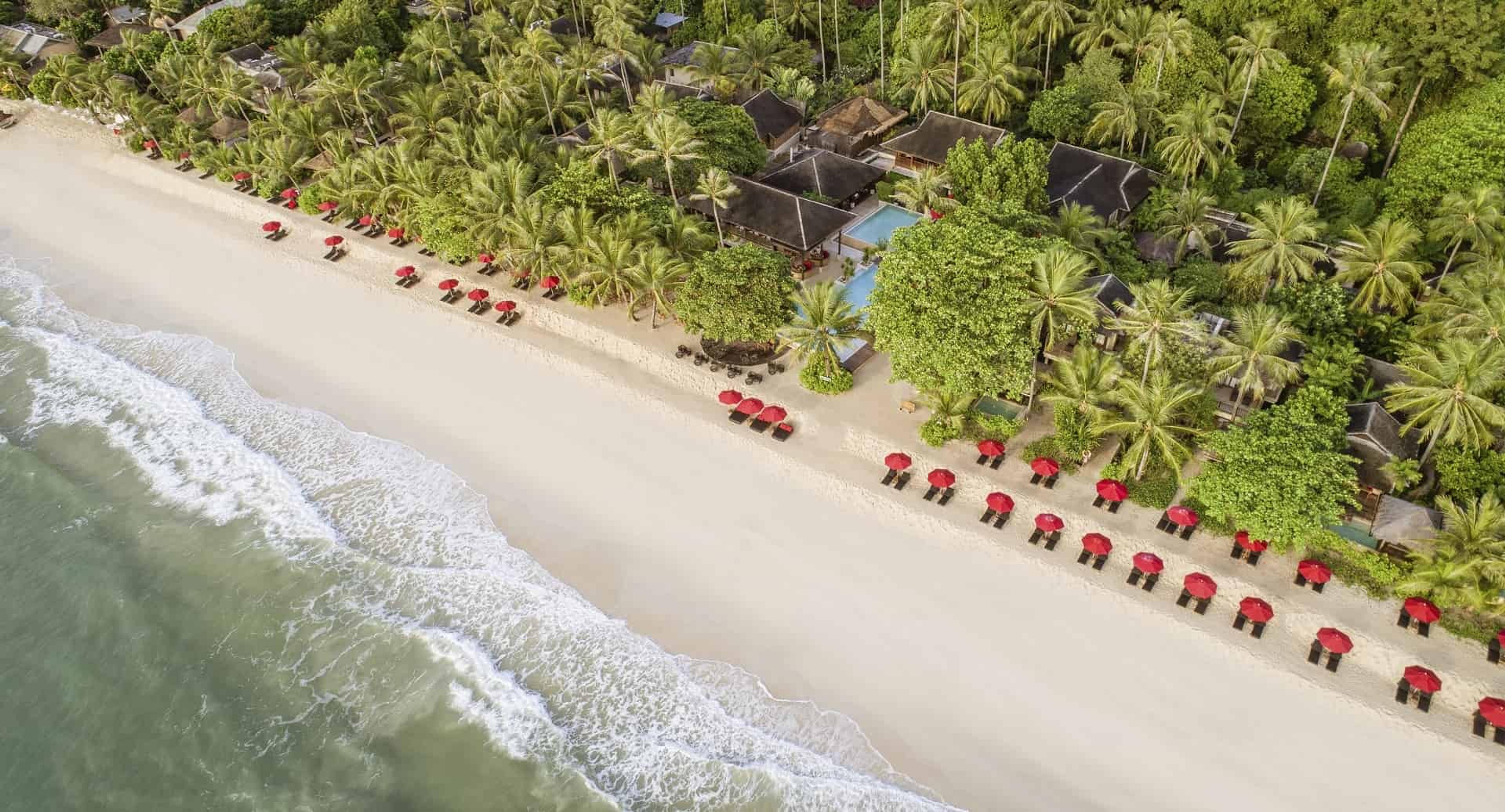 An overhead view of a white sand beach with a line of red umbrellas, waves breaking along the shore, and a pool and thatched roofs set among the palms behind it.