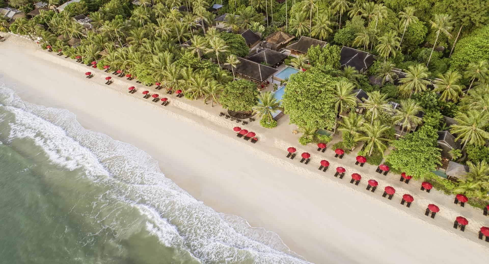 An overhead view of a white sand beach with a line of red umbrellas, waves breaking along the shore, and a pool and thatched roofs set among the palms behind it.