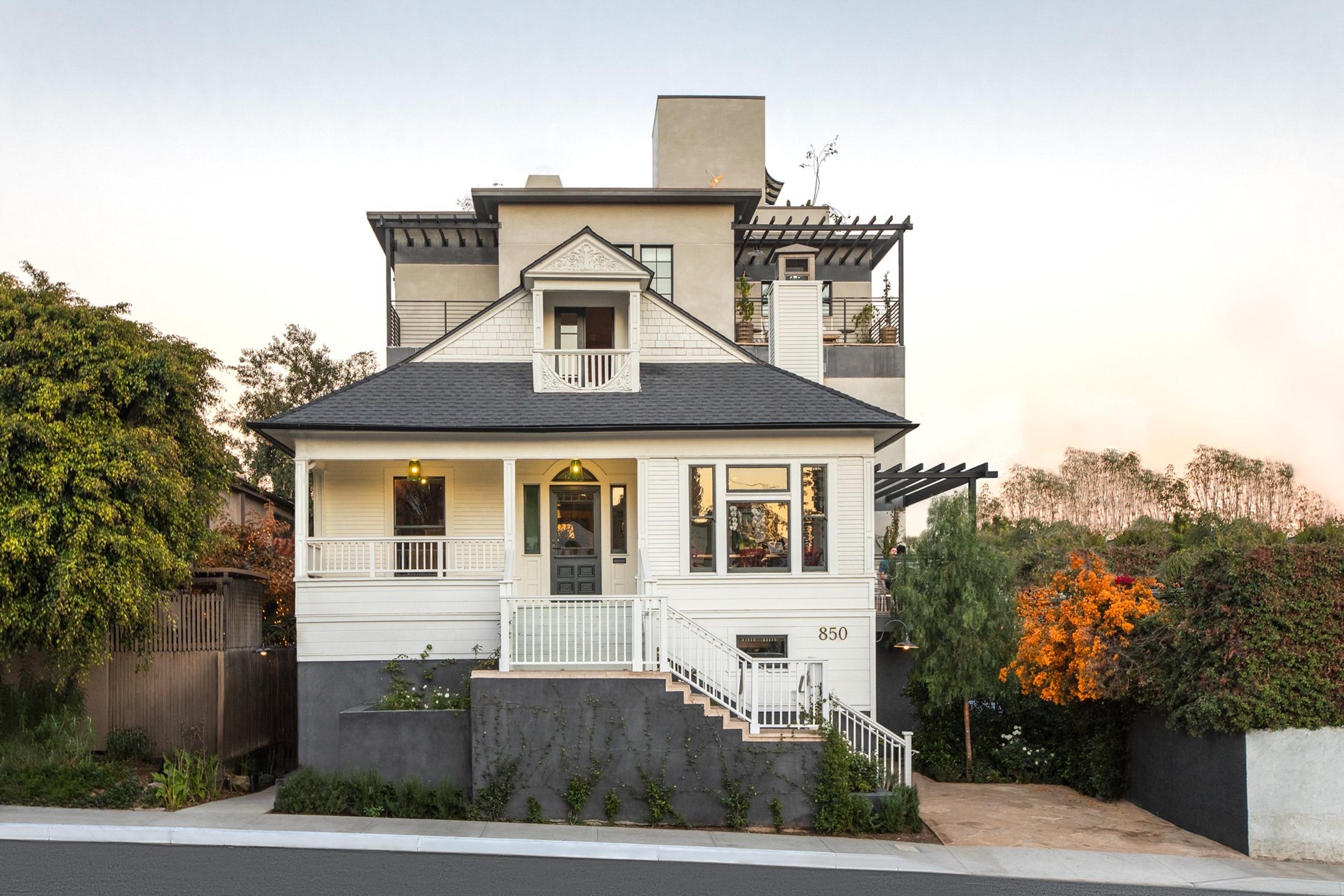 A two-storey Craftsman house with white clapboard siding, a dark gabled roof, and a modern stucco addition rising above it, photographed at dusk.