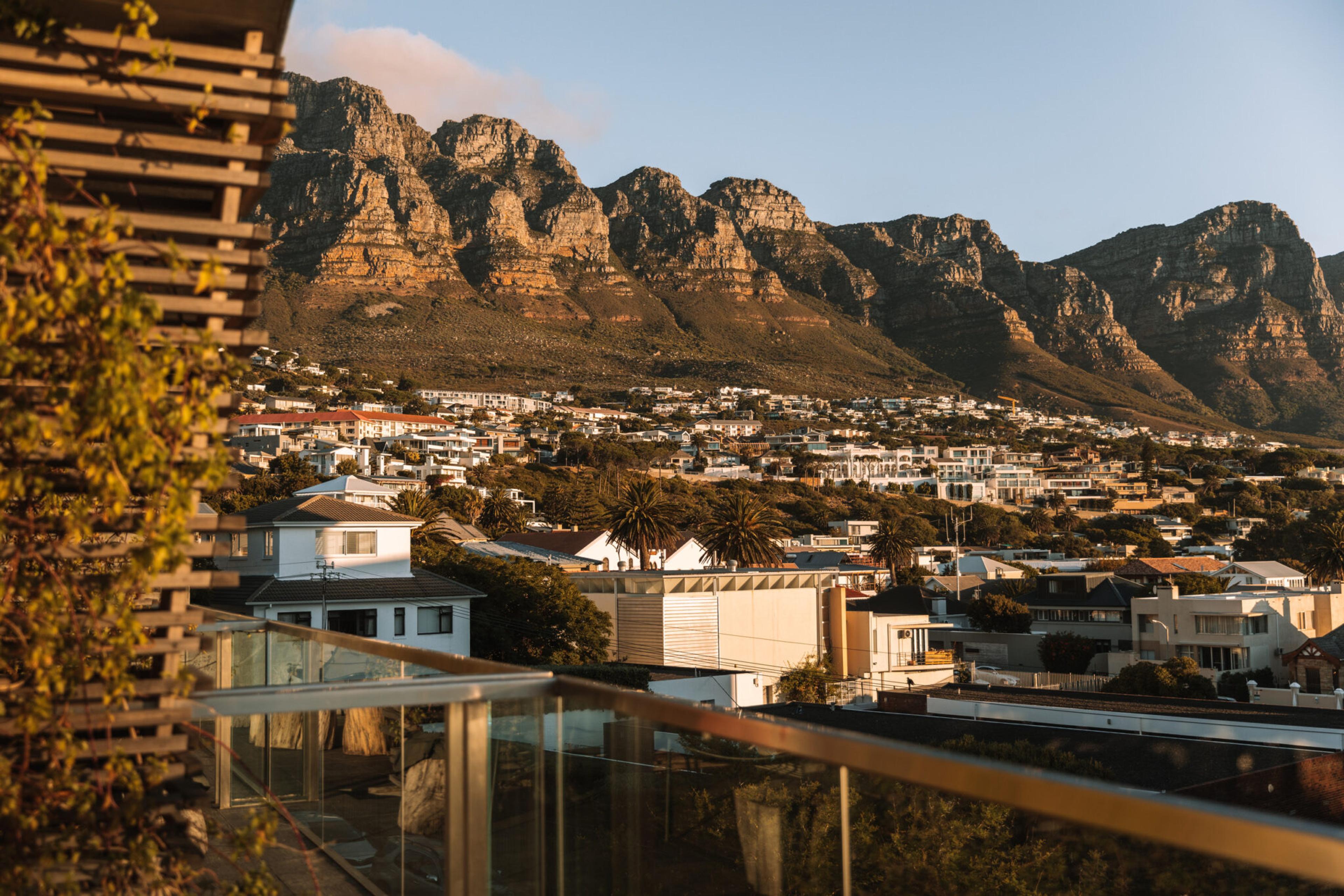 A timber-slatted rooftop terrace with a glass balustrade, looking out over suburban rooftops and palm trees to the Table Mountain range at golden hour.