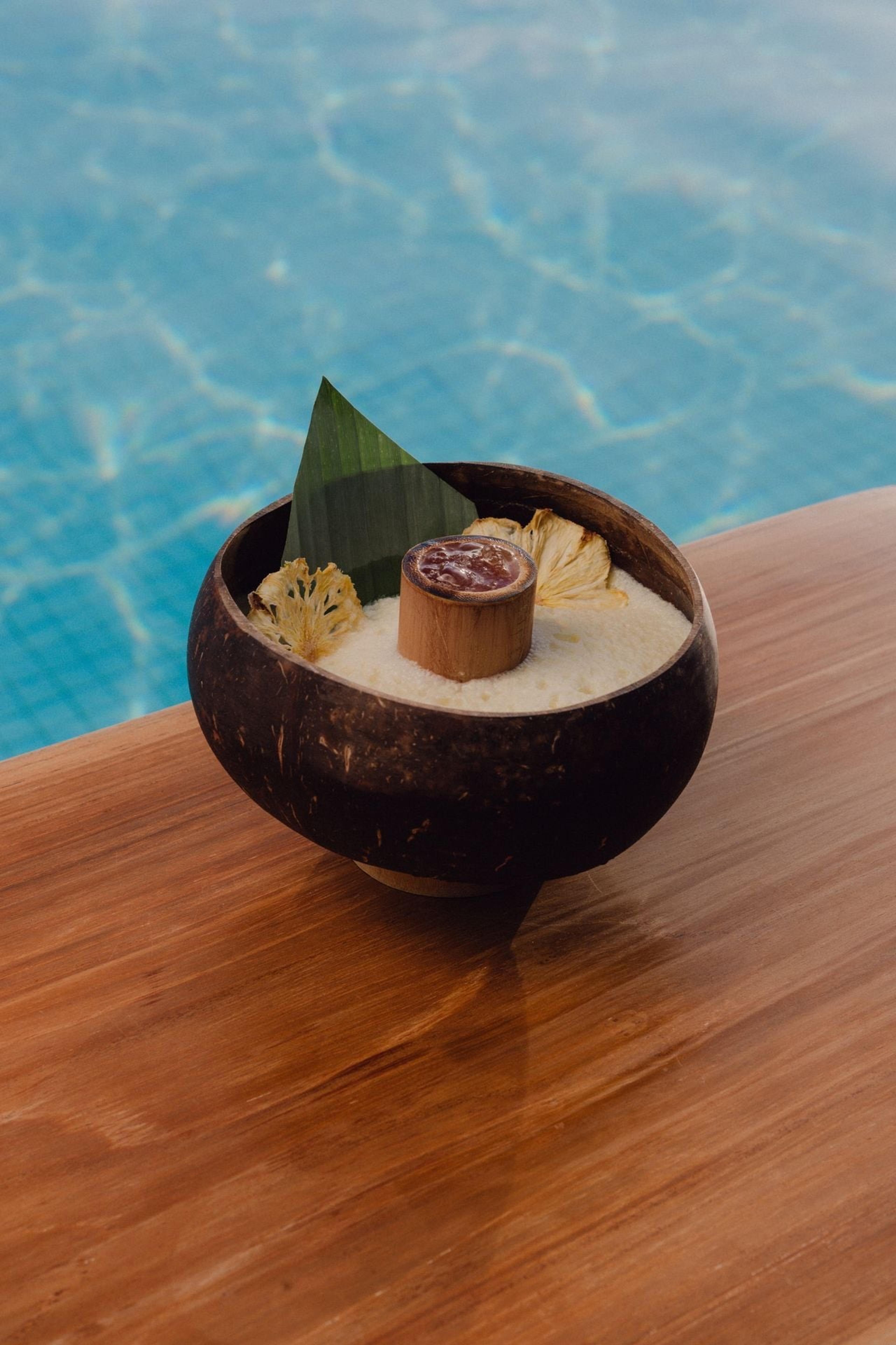 A dessert served in a coconut shell with a dried pineapple flower and a leaf garnish, set on a wooden ledge beside a pool.