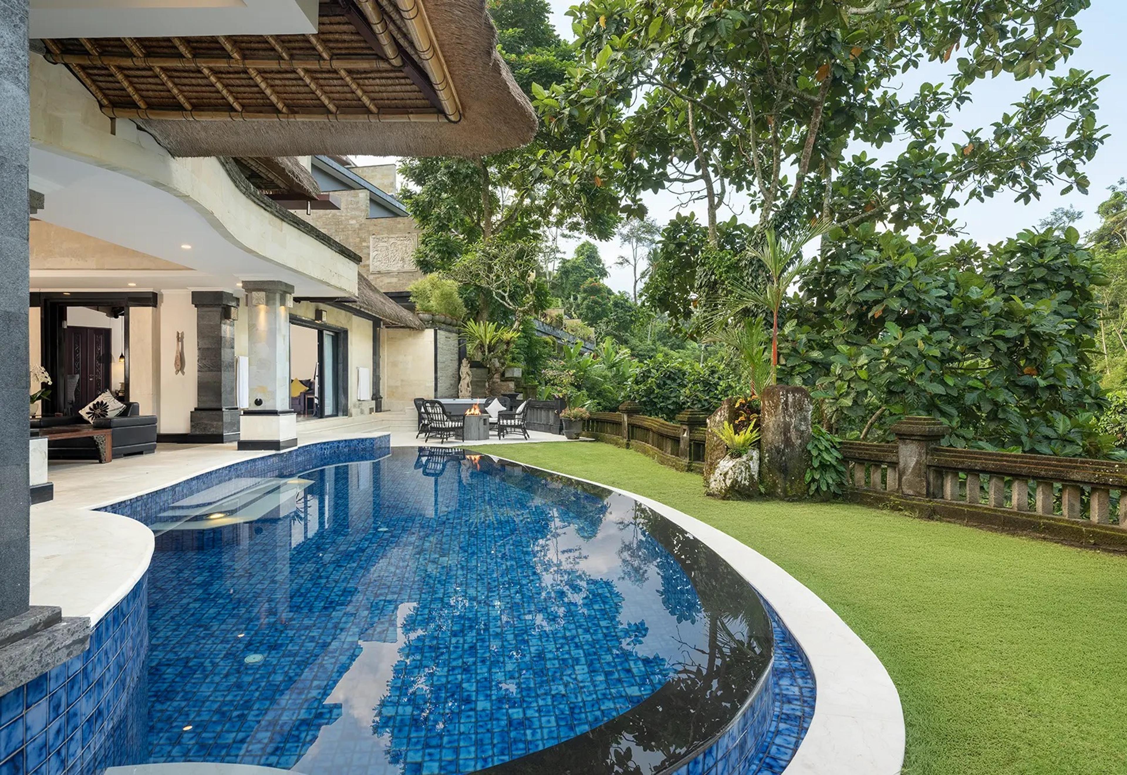 A curved blue mosaic pool runs along a two storey stone villa, with a lawn and trees on the other side.
