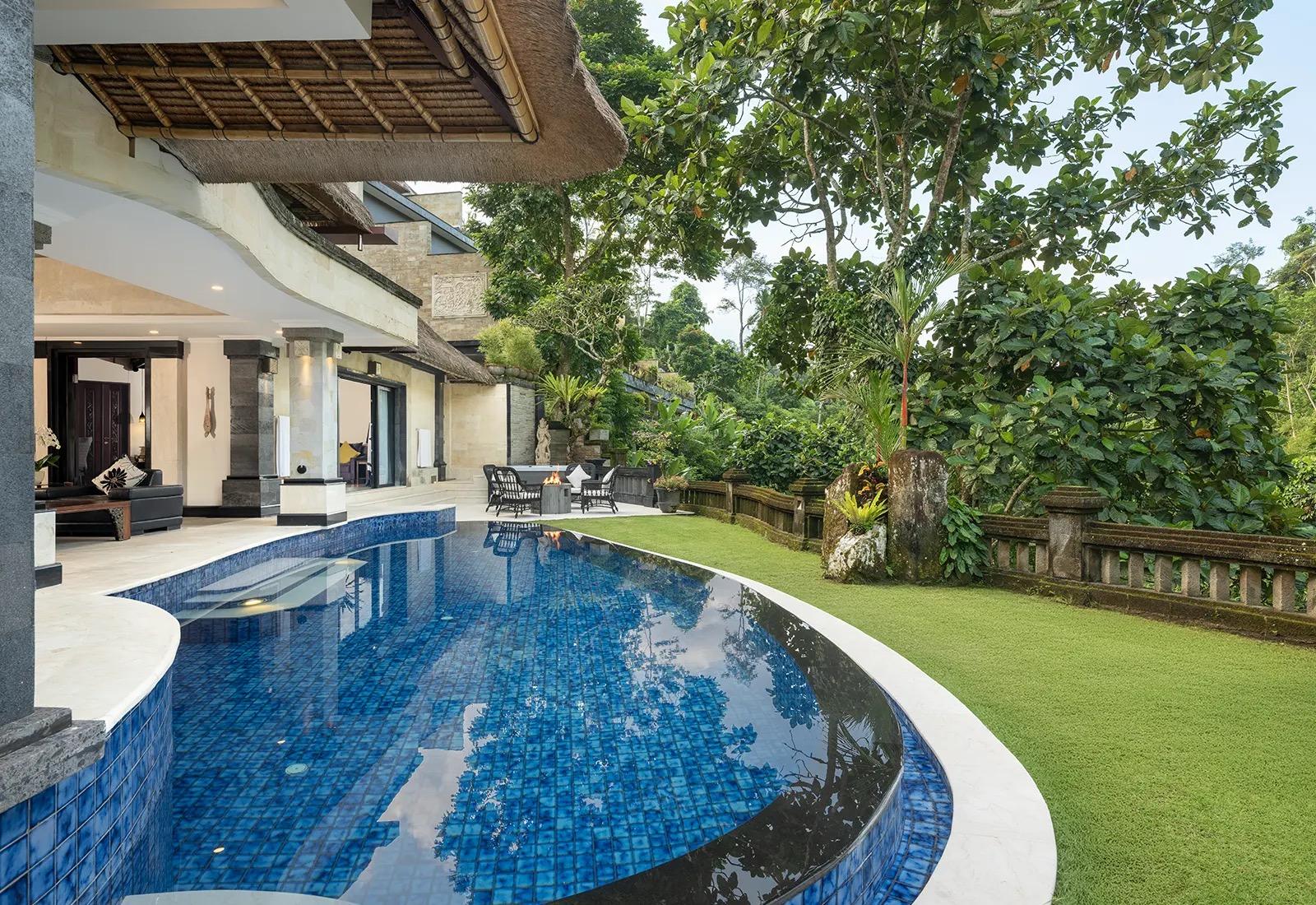 A curved blue mosaic pool runs along a two storey stone villa, with a lawn and trees on the other side.