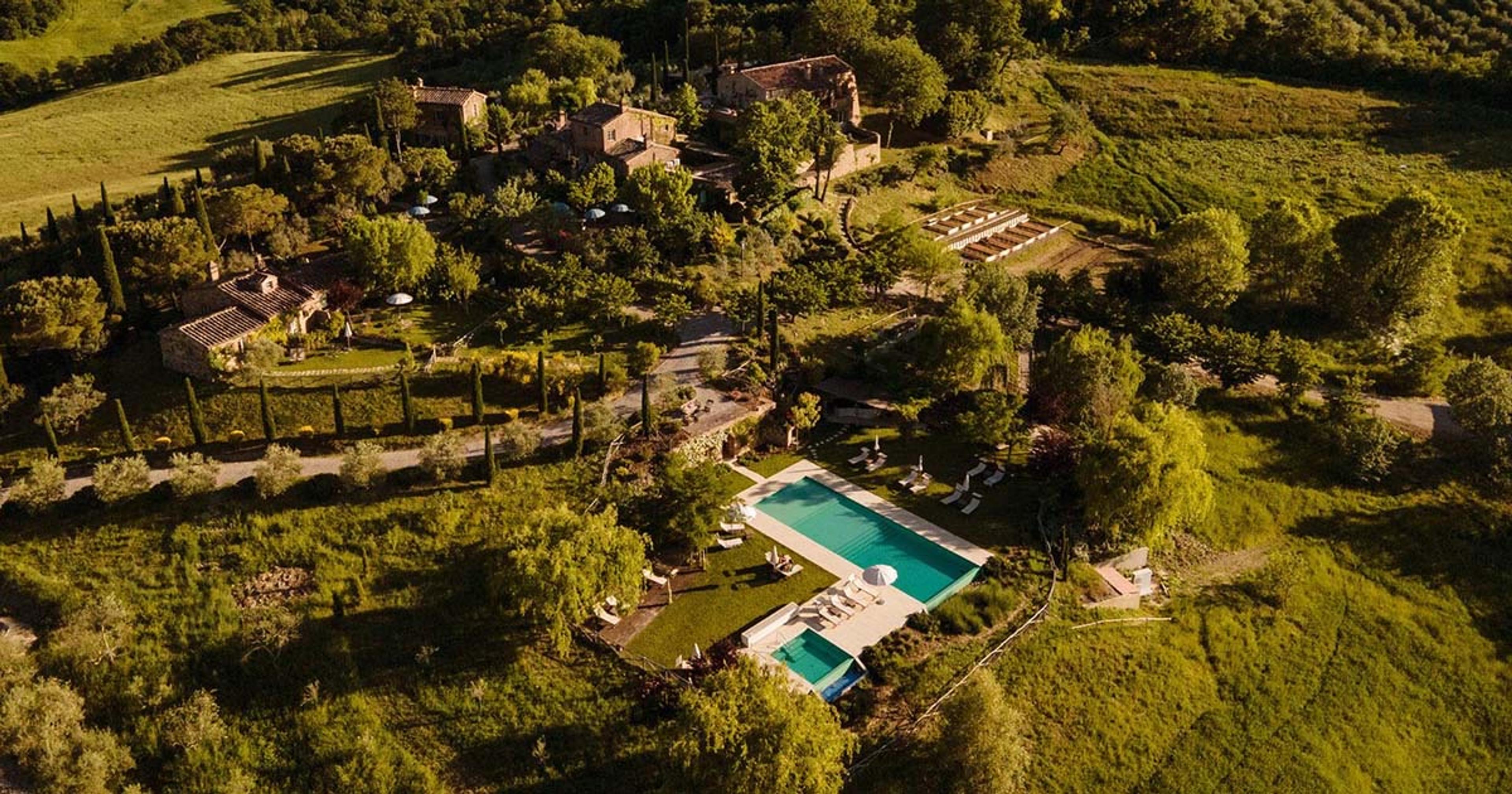 A stone farmhouse estate seen from above at golden hour, with cypress-lined paths, gardens and a swimming pool set among rolling hills.