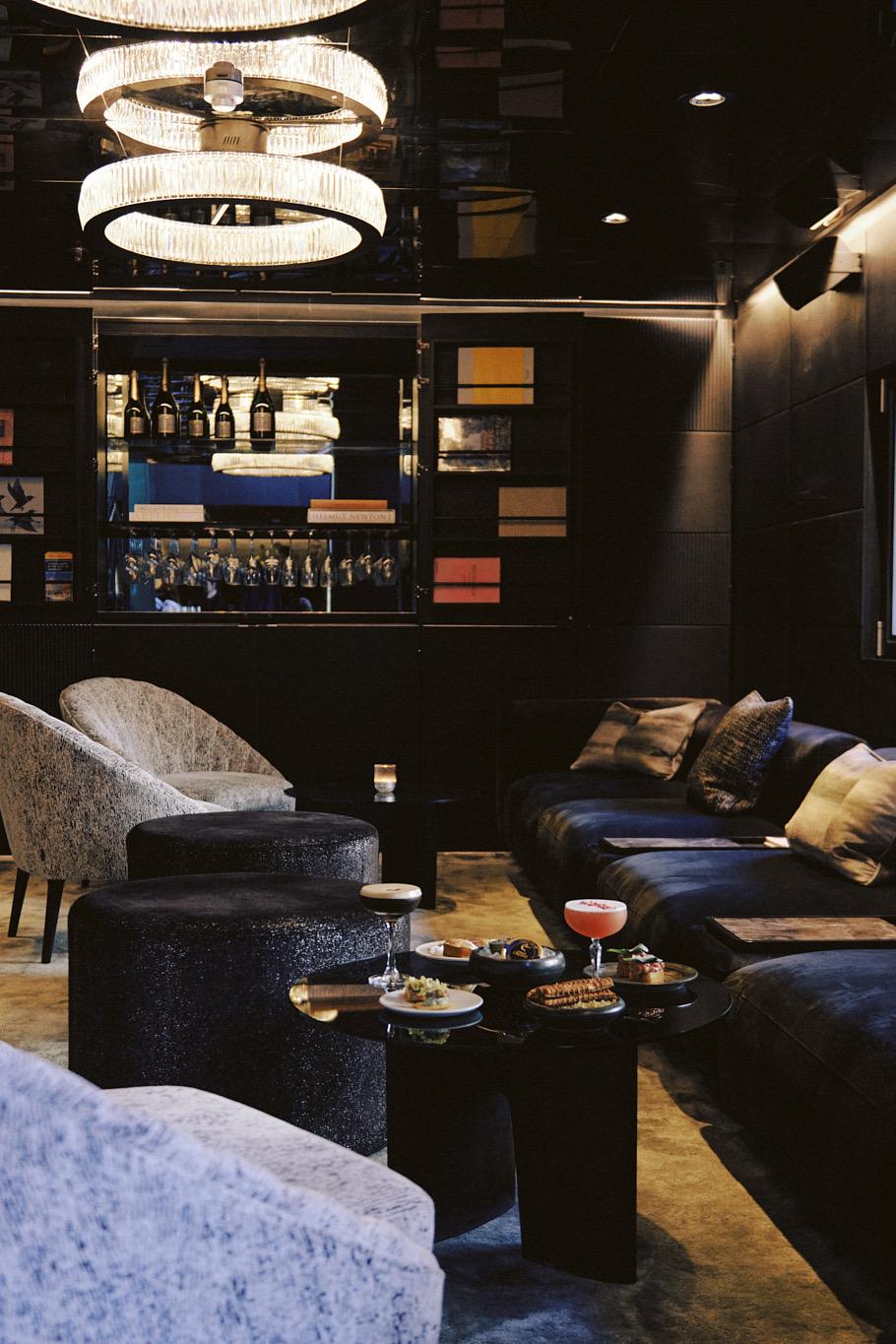 A dark lounge with armchairs, a low table set with cocktails and small plates, a sofa with cushions and a shelved bar in the background, lit by a chandelier.