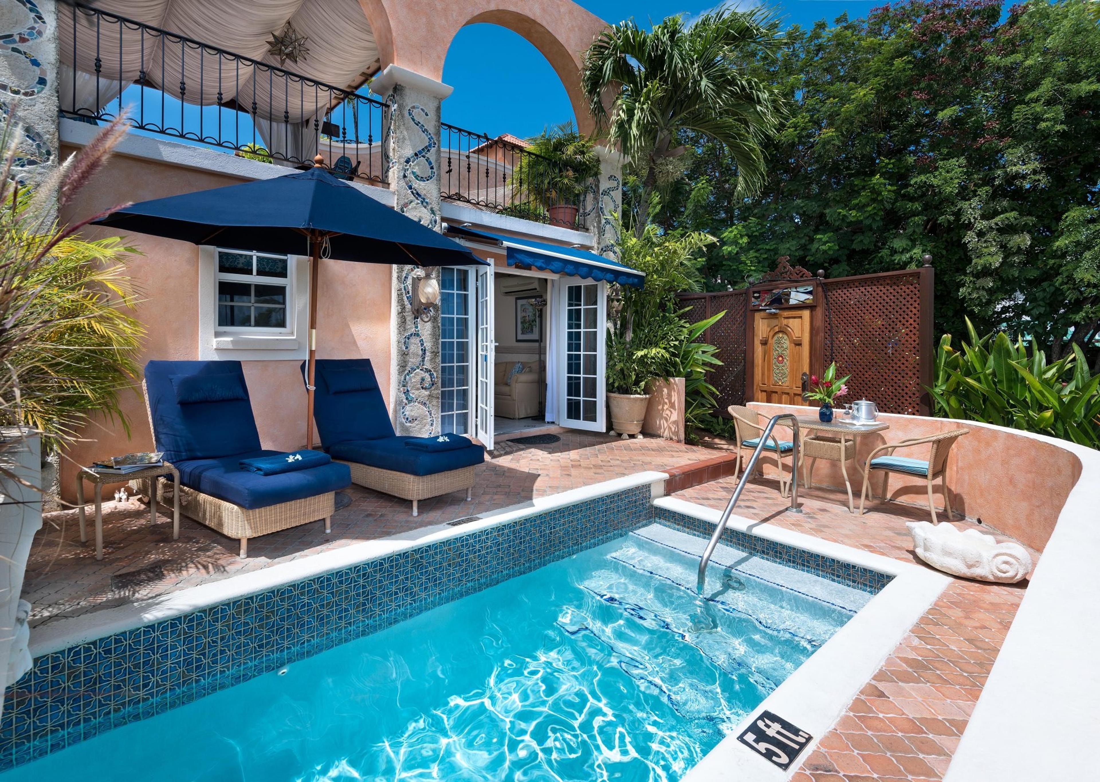A private plunge pool beside a terrace with two blue loungers, a navy umbrella, and an arched balcony with mosaic pillars above.