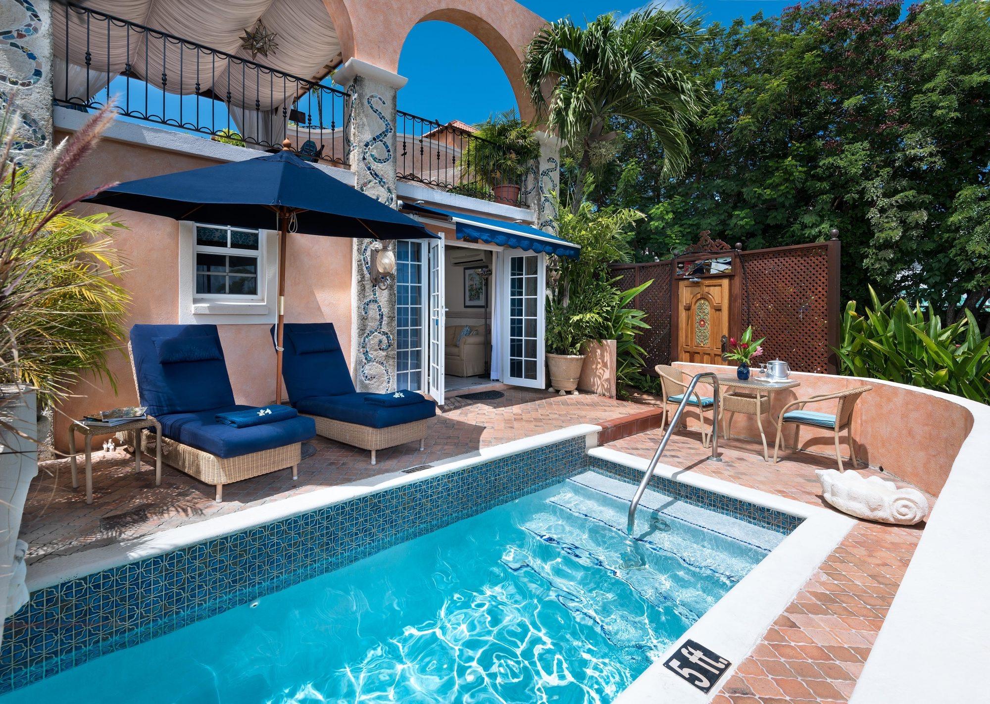 A private plunge pool beside a terrace with two blue loungers, a navy umbrella, and an arched balcony with mosaic pillars above.
