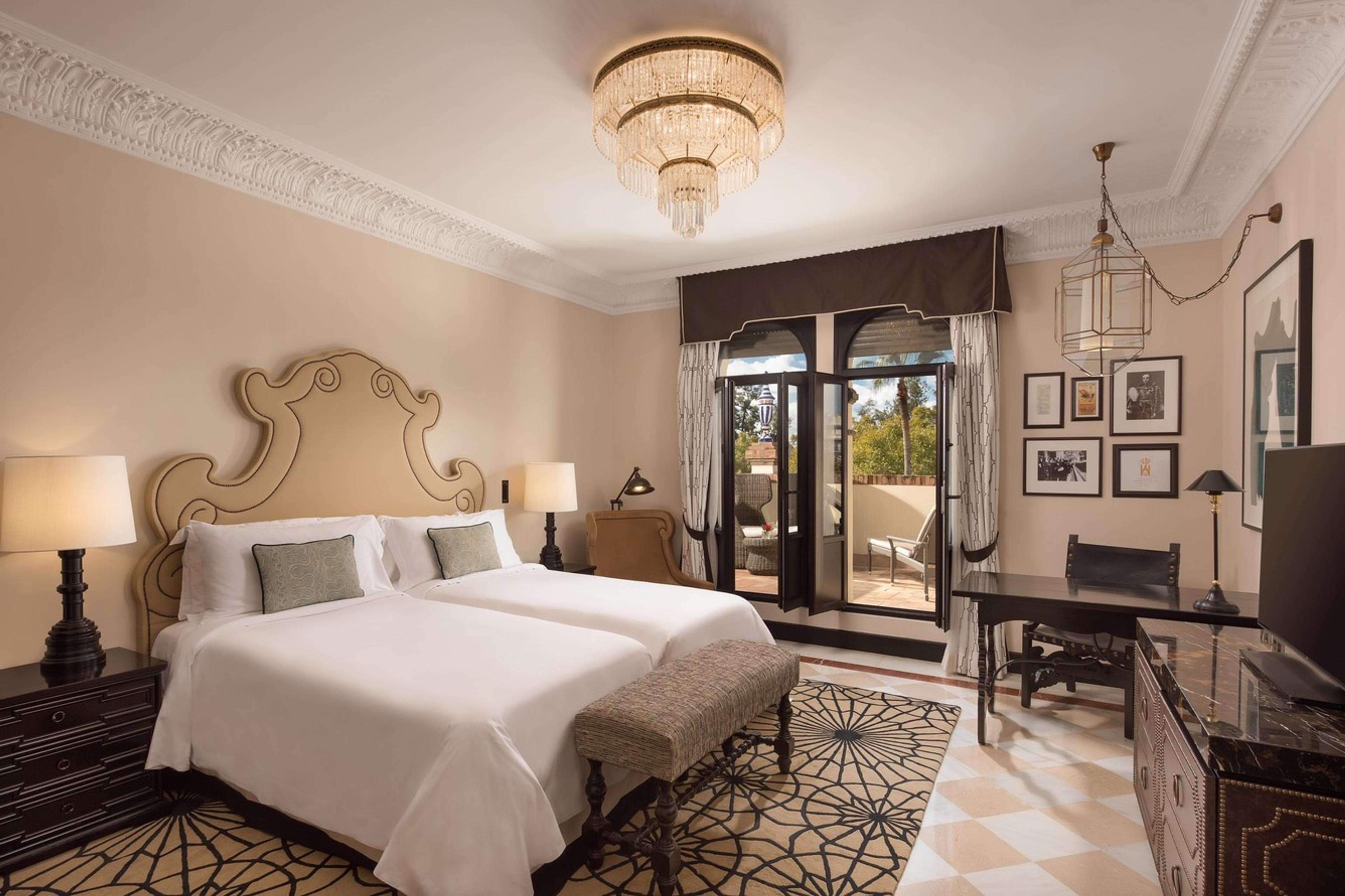A suite with twin beds under a gold-scrolled wood headboard, bedside lamps, a wall of framed photographs, a writing desk and television, and open balcony doors leading to an outdoor seating area.