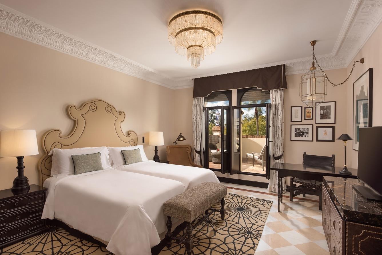 A suite with twin beds under a gold-scrolled wood headboard, bedside lamps, a wall of framed photographs, a writing desk and television, and open balcony doors leading to an outdoor seating area.
