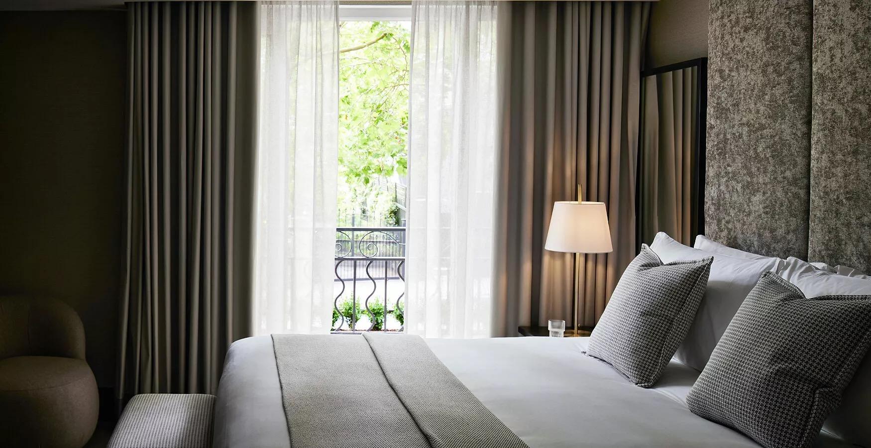 A bed dressed in white and grey linen facing a glass door with sheer curtains open to a wrought iron balcony and a tree outside.