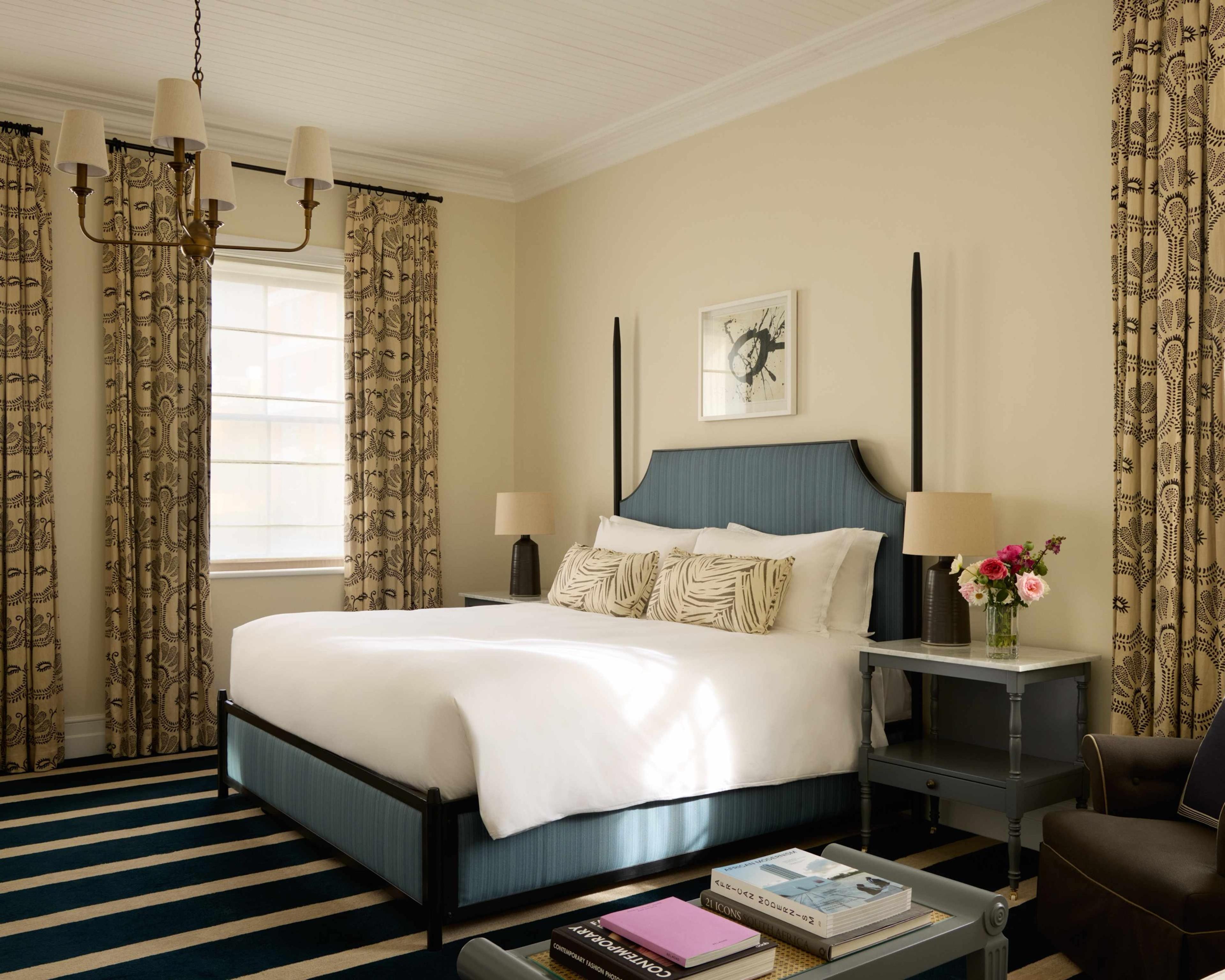 A bedroom with a large bed under a blue upholstered headboard, a window with patterned curtains, a striped rug, and a chandelier above.