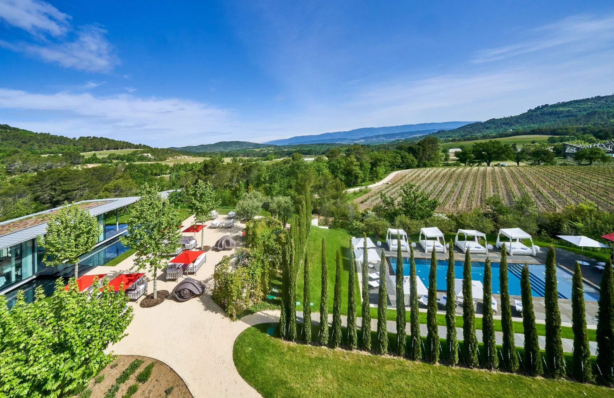 An aerial view of a glass-walled building beside a landscaped pool deck with white loungers, a row of cypress trees, dark sculptural forms on the lawn, and vineyards and hills beyond.