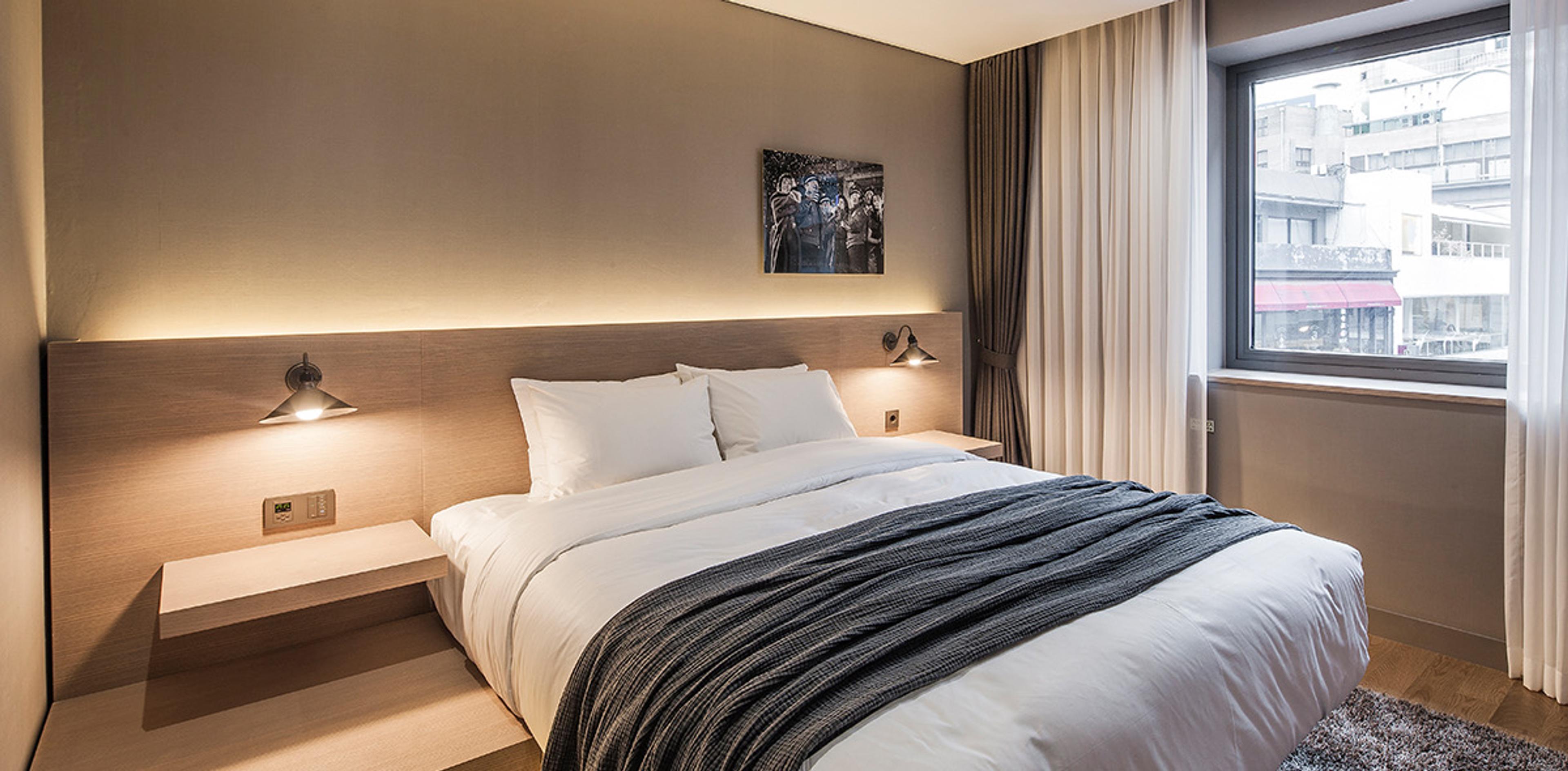 A hotel bedroom with a bed against a tan wall, a small black and white photograph above the headboard, and a large bright window looking over the rooftops and buildings outside.