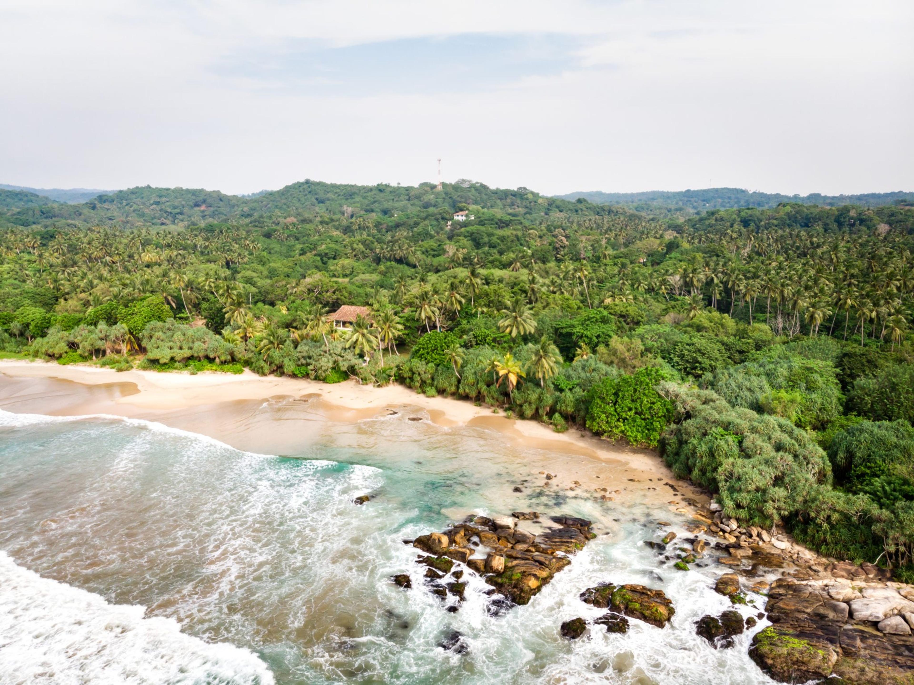A white sand beach curves around a rocky point, with turquoise surf breaking over the rocks and a palm grove and a red tiled roof behind it, seen from above.