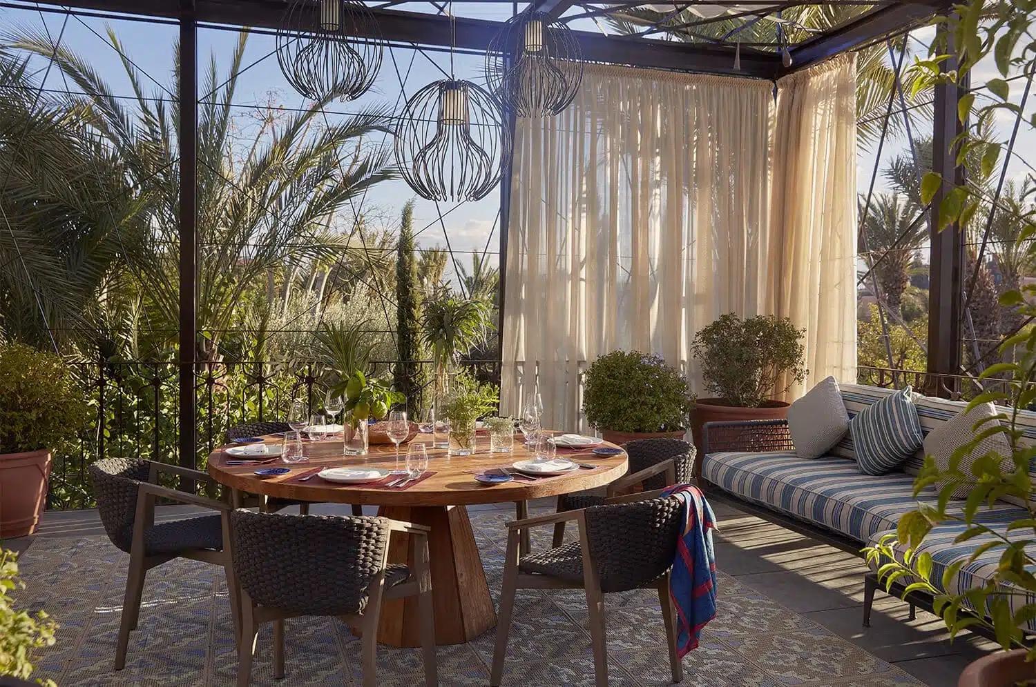 A covered terrace with a round table set for dinner, sheer curtains and wire pendant lights, surrounded by palm trees.