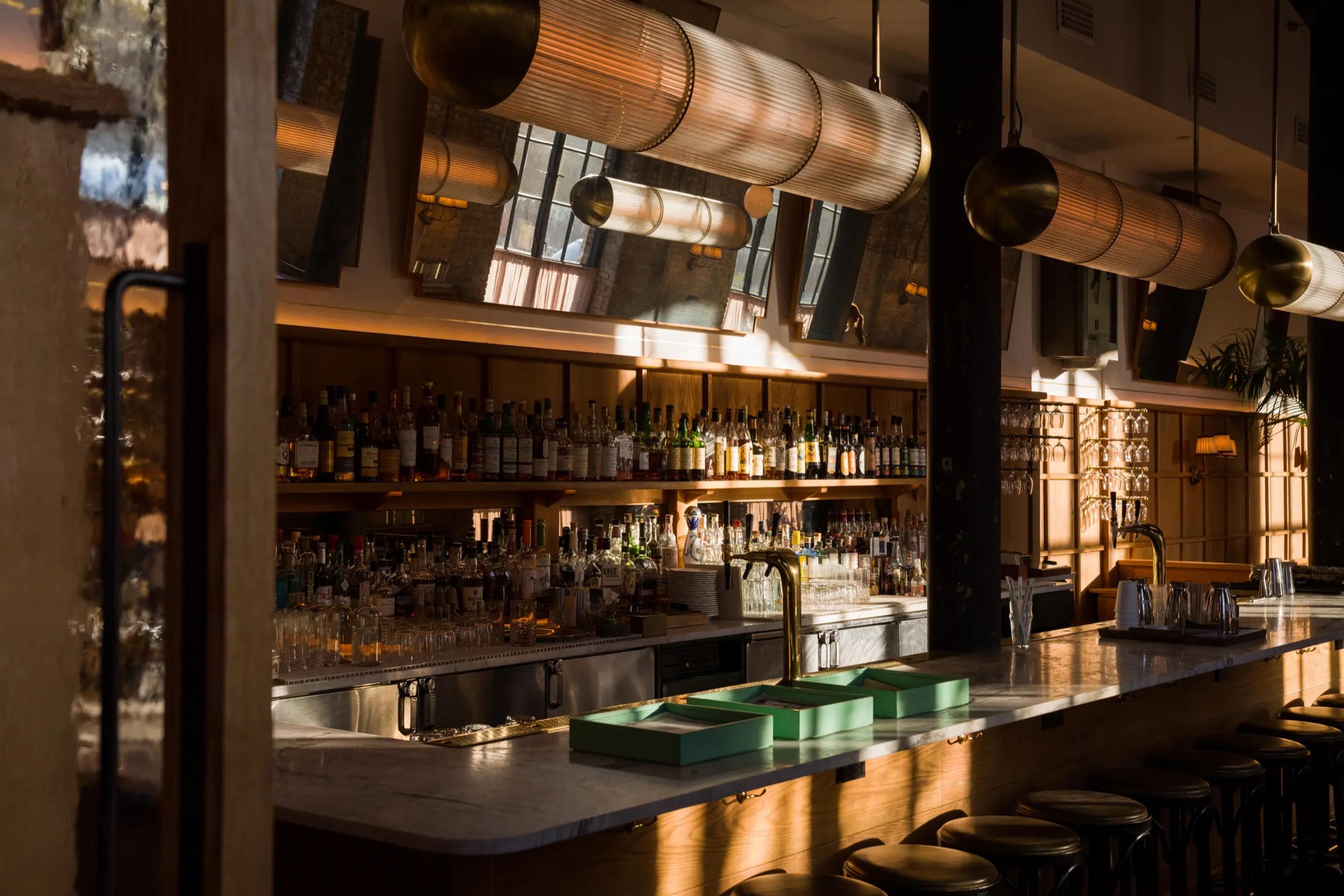 A long bar lined with liquor bottles and glassware under cylindrical glass pendant lights, with empty stools along the counter.