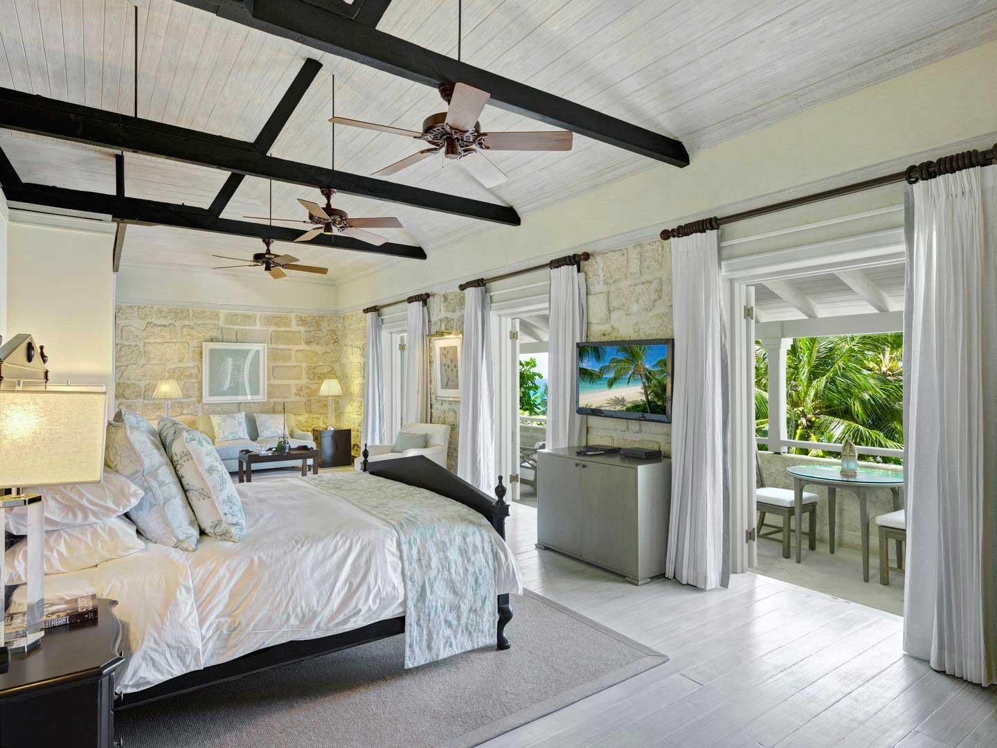 A bedroom under a white beamed ceiling with ceiling fans, a dark four-poster bed, coral-stone walls and doors open to a balcony