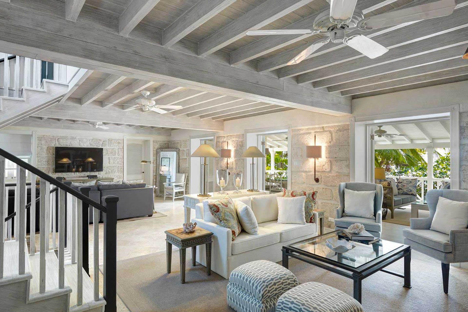 An open-plan living room under a wood-beamed ceiling, with a grey sofa, armchairs, a glass coffee table, and a staircase to one side.