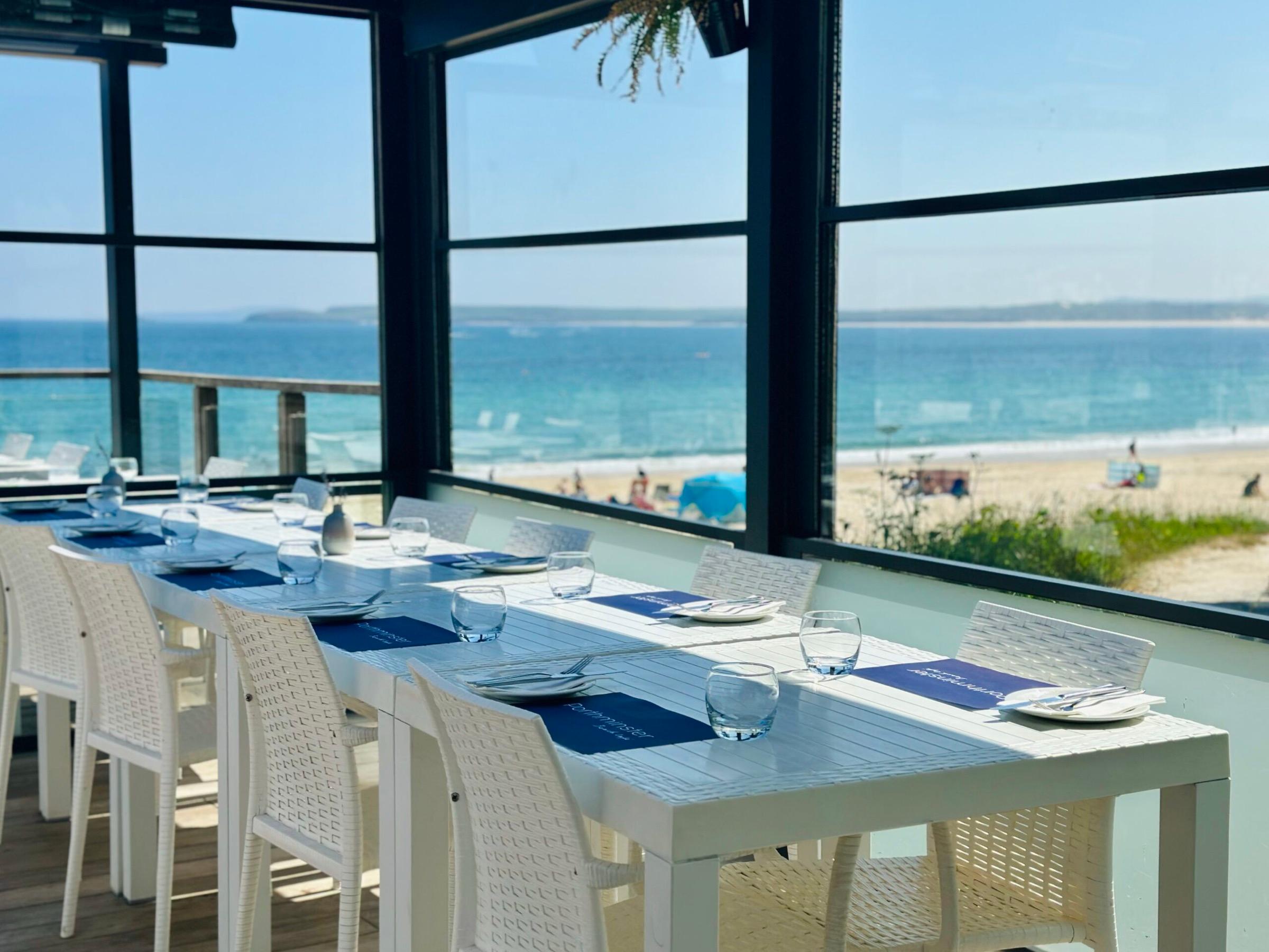 A dining room walled in glass, tables laid in white and blue, looking straight out over the sand and sea at St Ives.