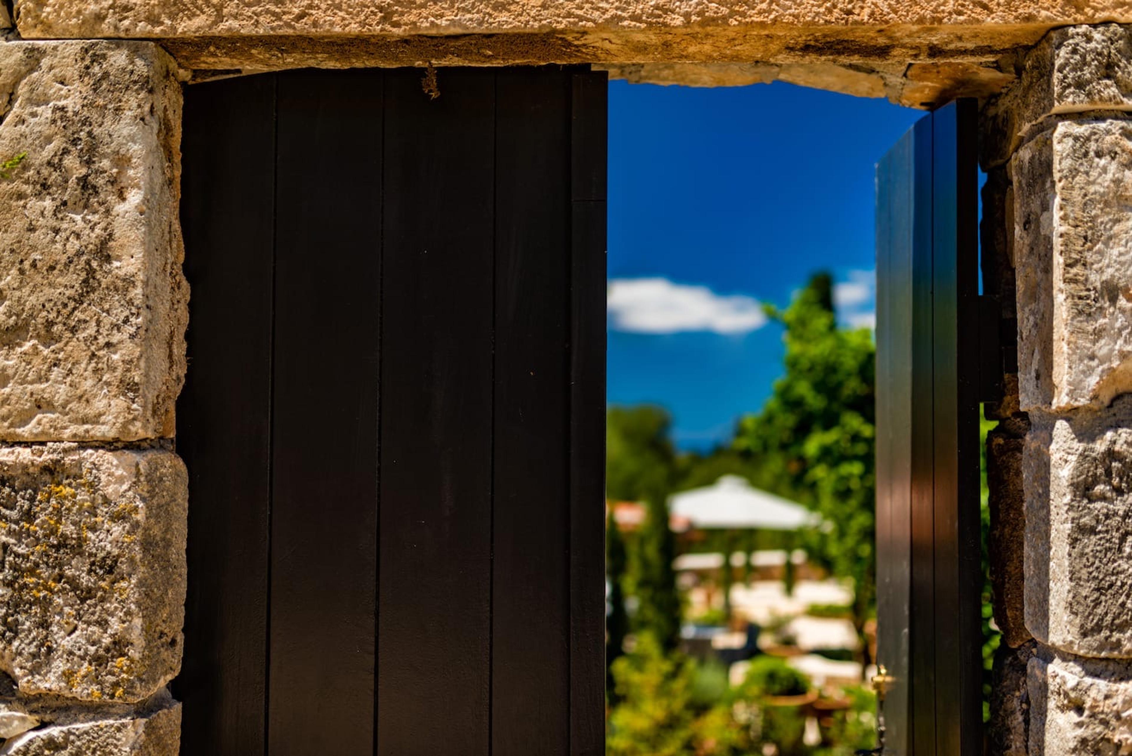 A black wooden door standing open in a stone archway, looking out to trees and sky with the foreground in soft focus.