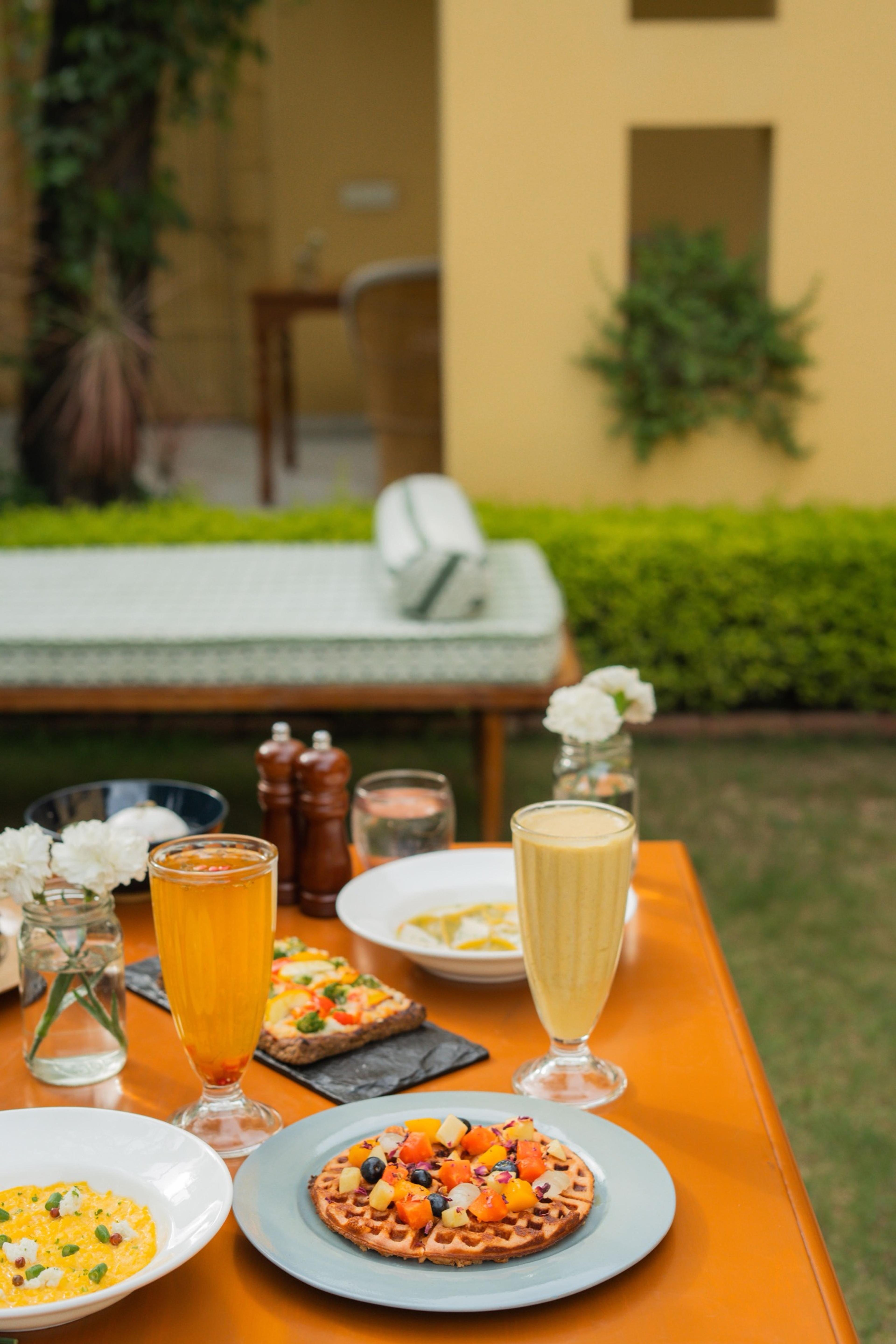 A waffle with fruit, a bowl of risotto, two glasses of juice and flowers laid on an orange outdoor table, with a daybed and the building out of focus behind.