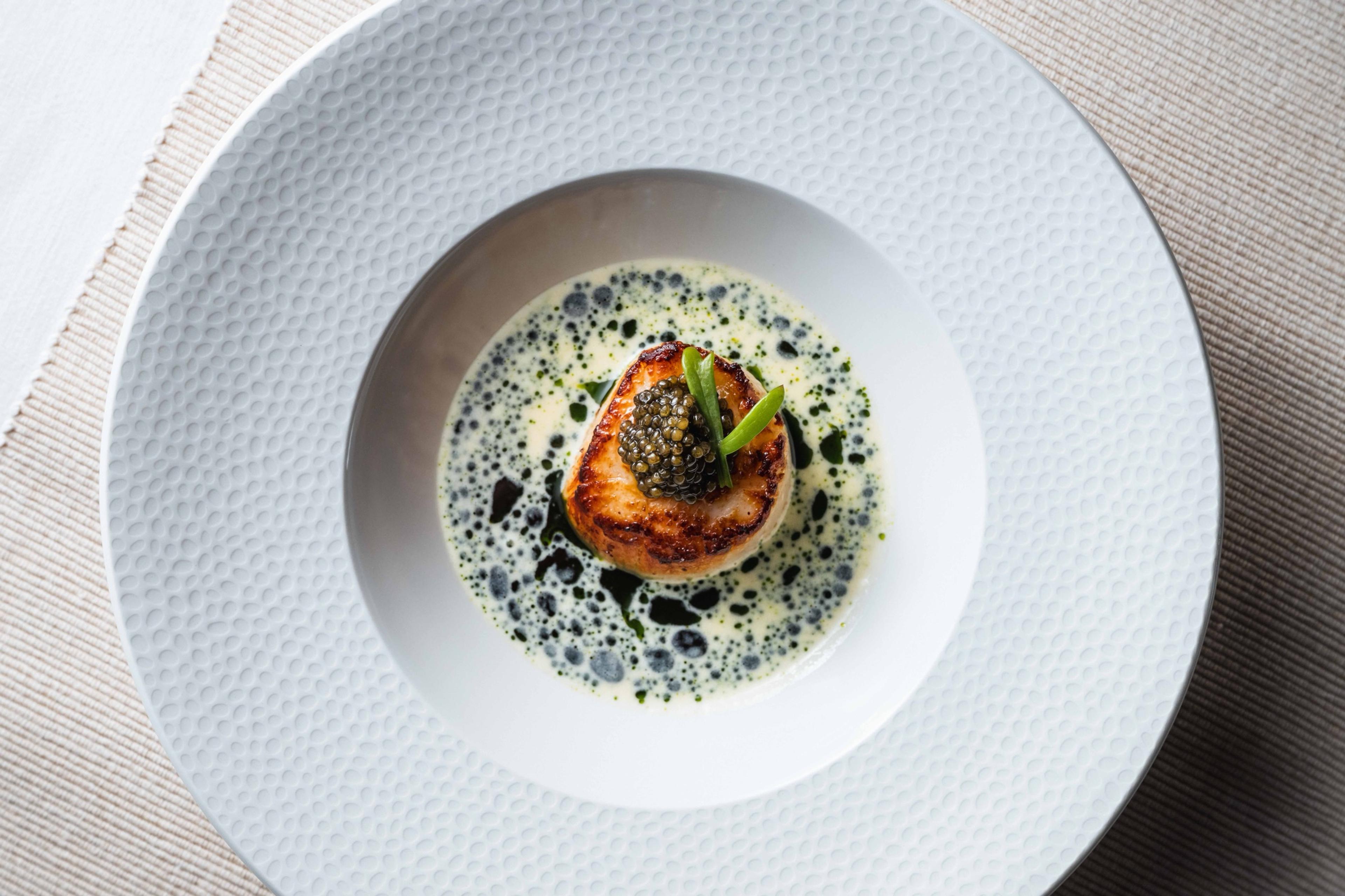 A seared scallop topped with caviar in a pale sauce with green herb oil, on a white textured plate.