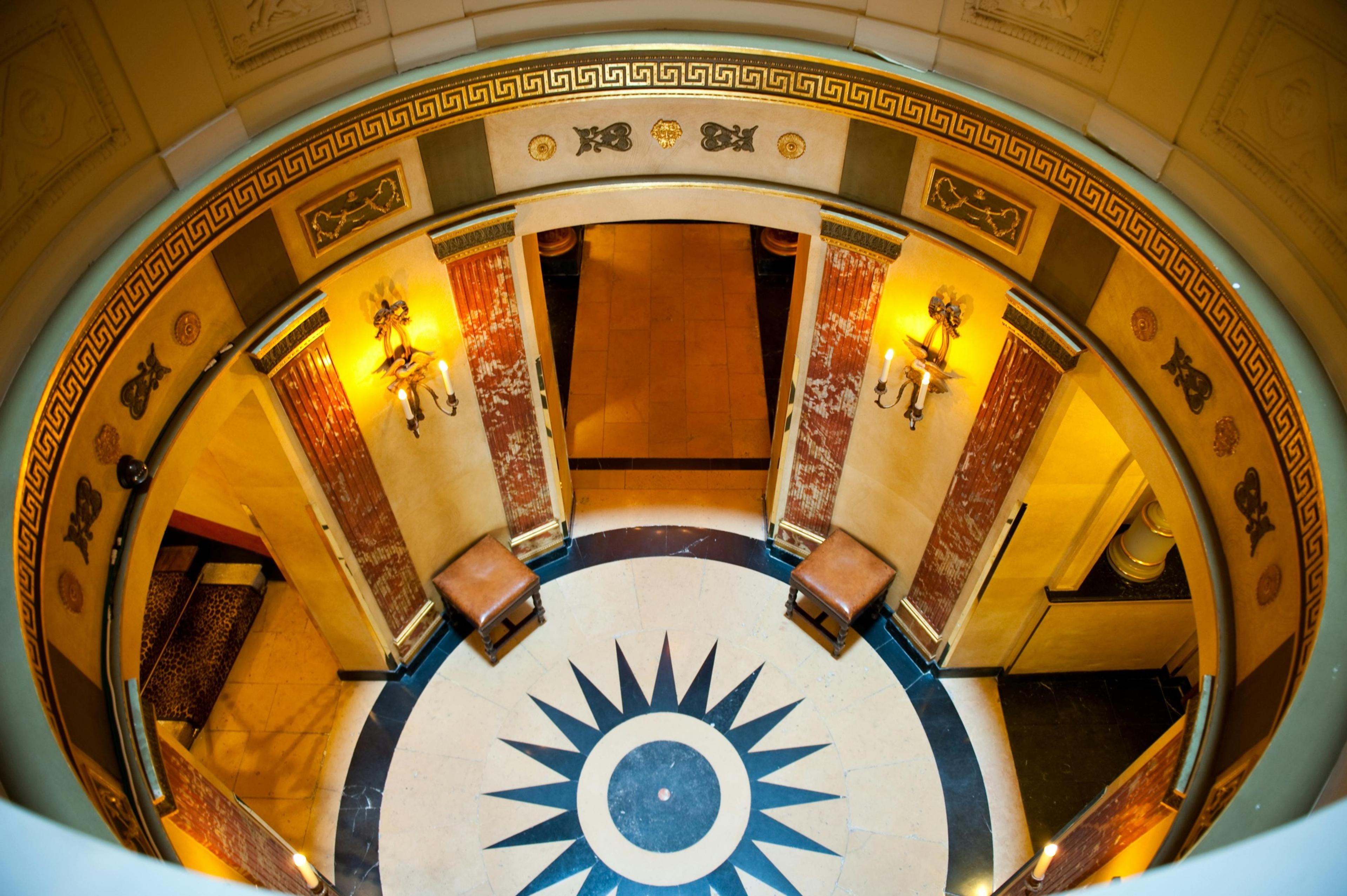 A fisheye view down a stairwell landing onto a black and white star-patterned marble floor, flanked by leather stools and wall sconces.