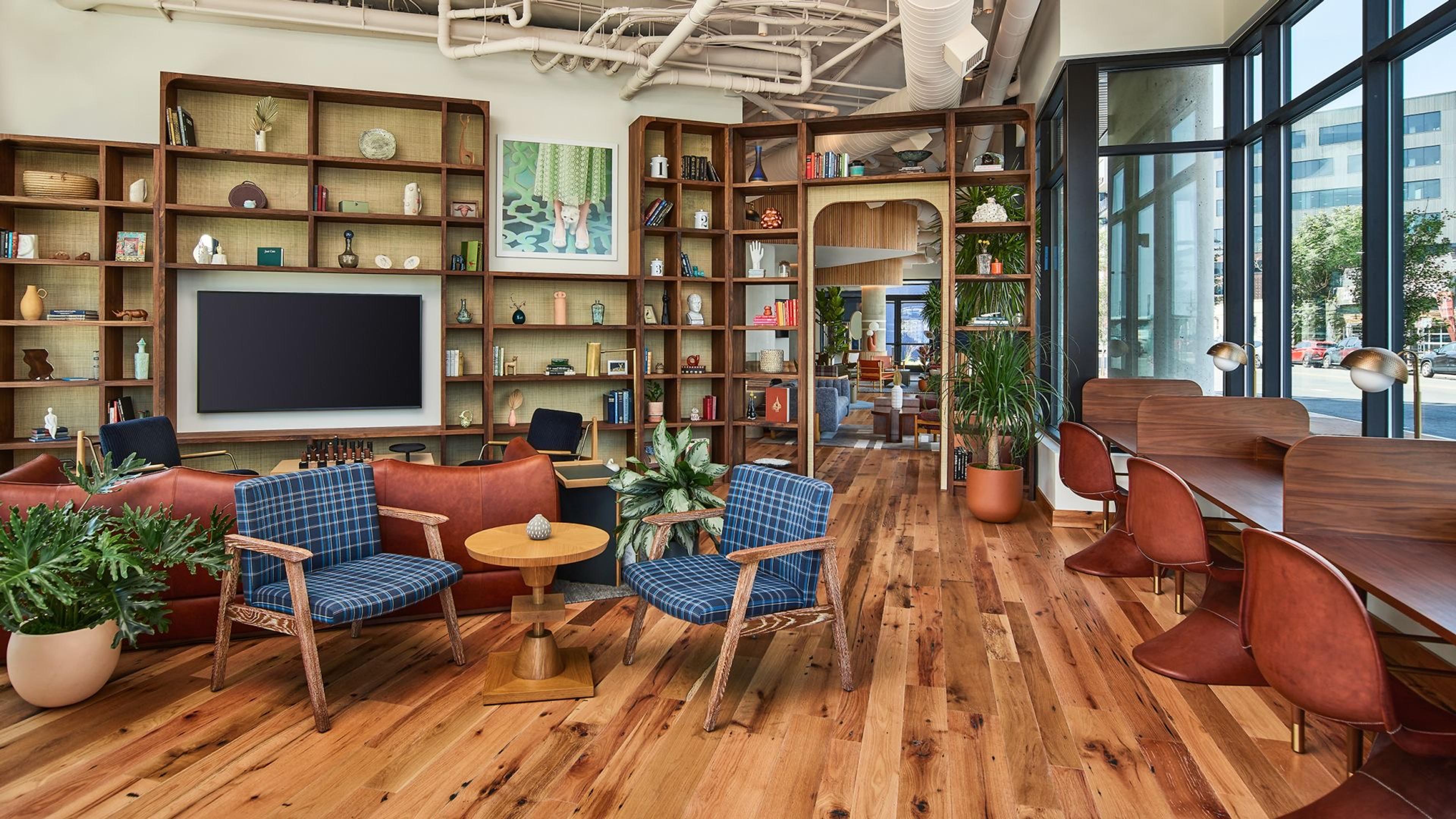 A hotel lounge with built-in wooden bookshelves filled with books and objects, a wall-mounted television, blue plaid armchairs and a long communal table by large windows.