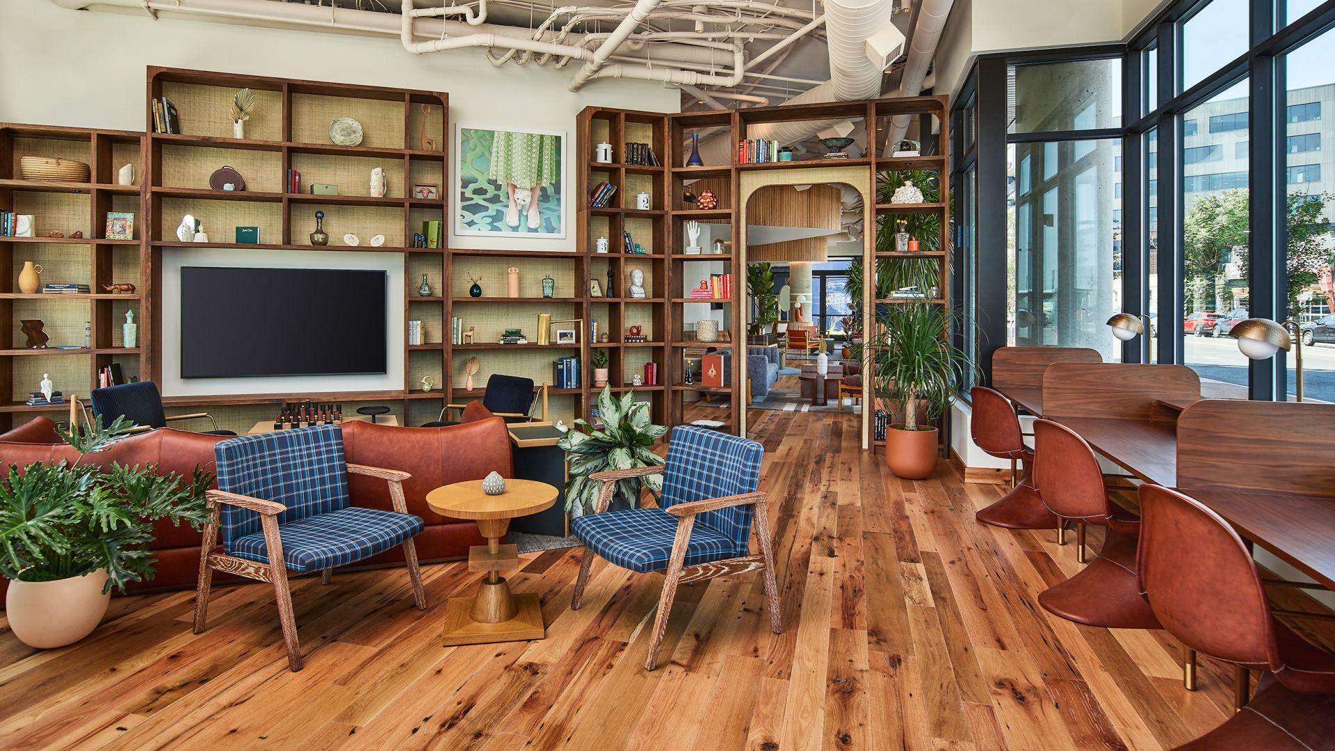 A hotel lounge with built-in wooden bookshelves filled with books and objects, a wall-mounted television, blue plaid armchairs and a long communal table by large windows.