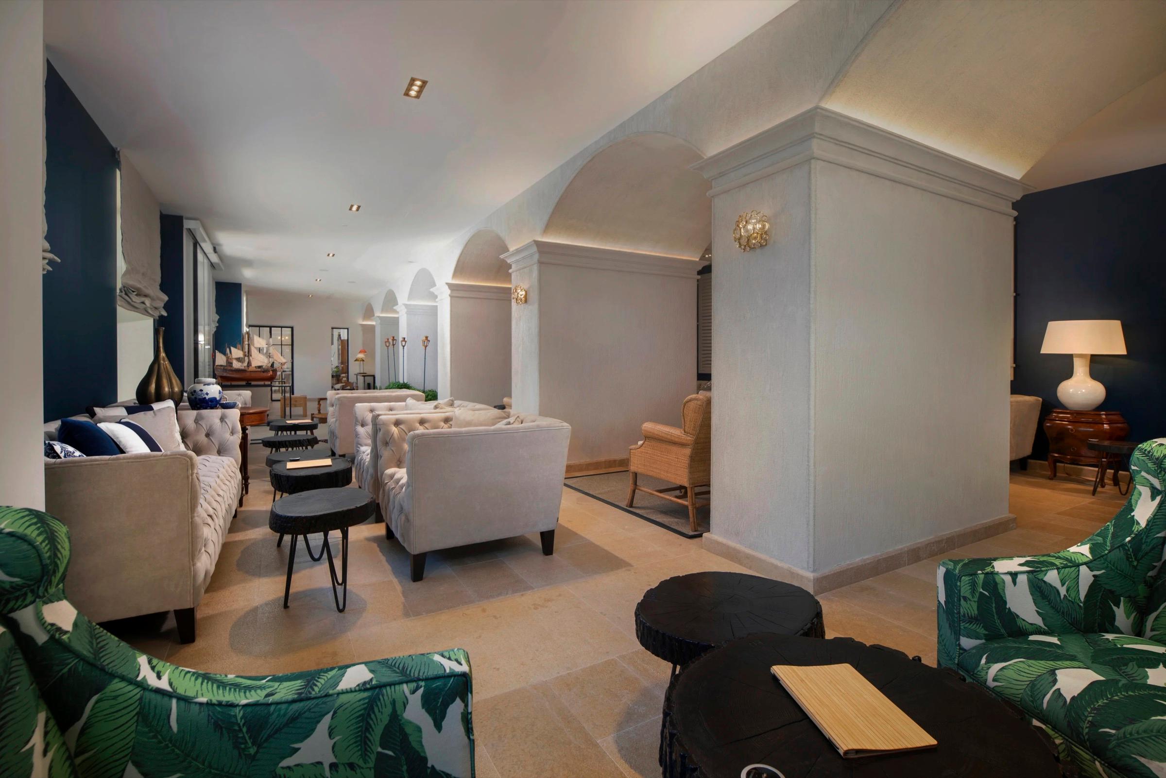 A hotel lounge with cream sofas, dark side tables, and two armchairs upholstered in a green palm-leaf print in the foreground.