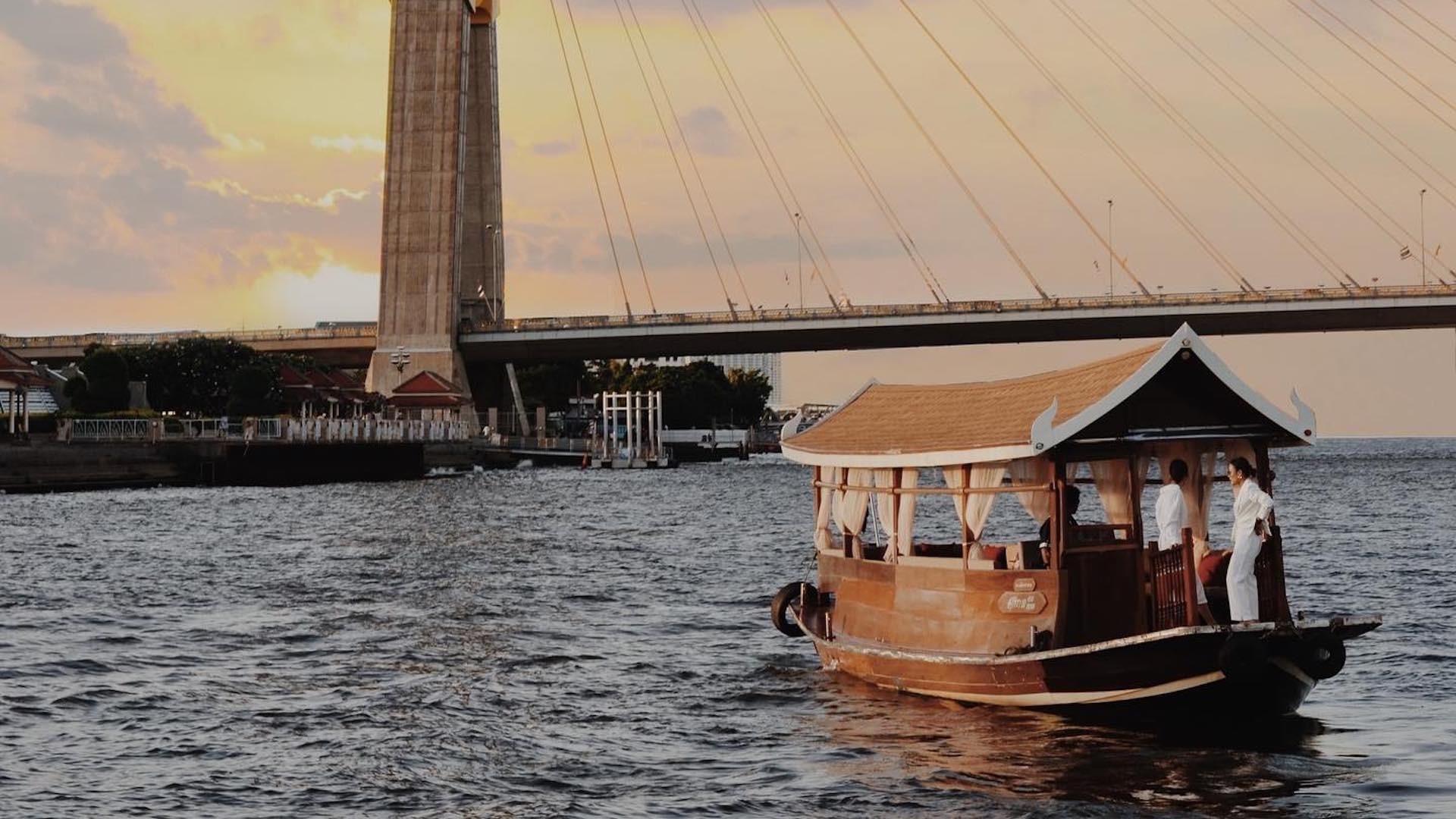 A traditional wooden river boat with a peaked roof carries a few people in white along the Chao Phraya at sunset, a cable-stayed bridge crossing the water behind it.