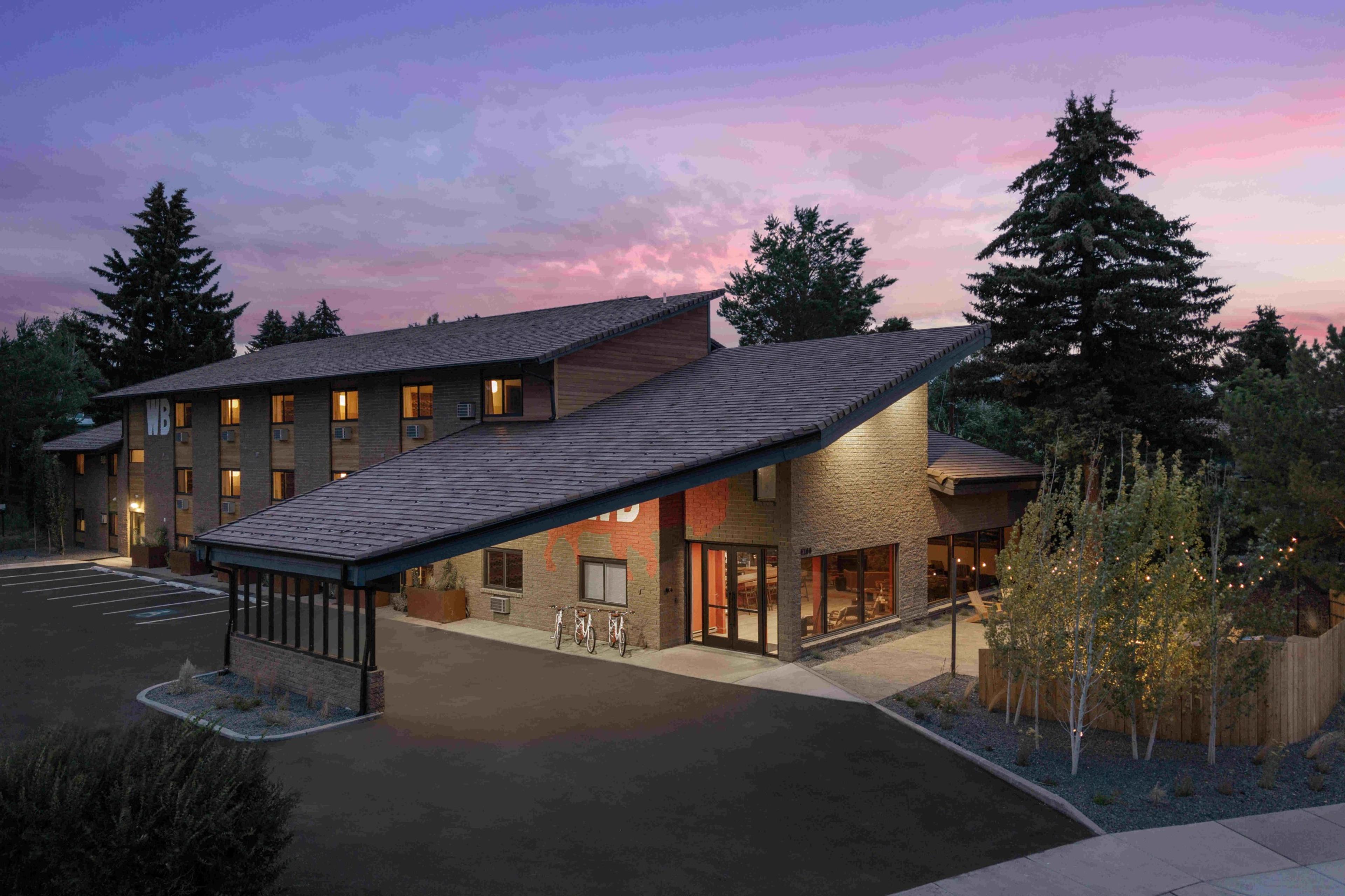 The hotel's exterior at dusk, its windows lit from inside, pine trees and a bicycle rack out front under a pink sky.