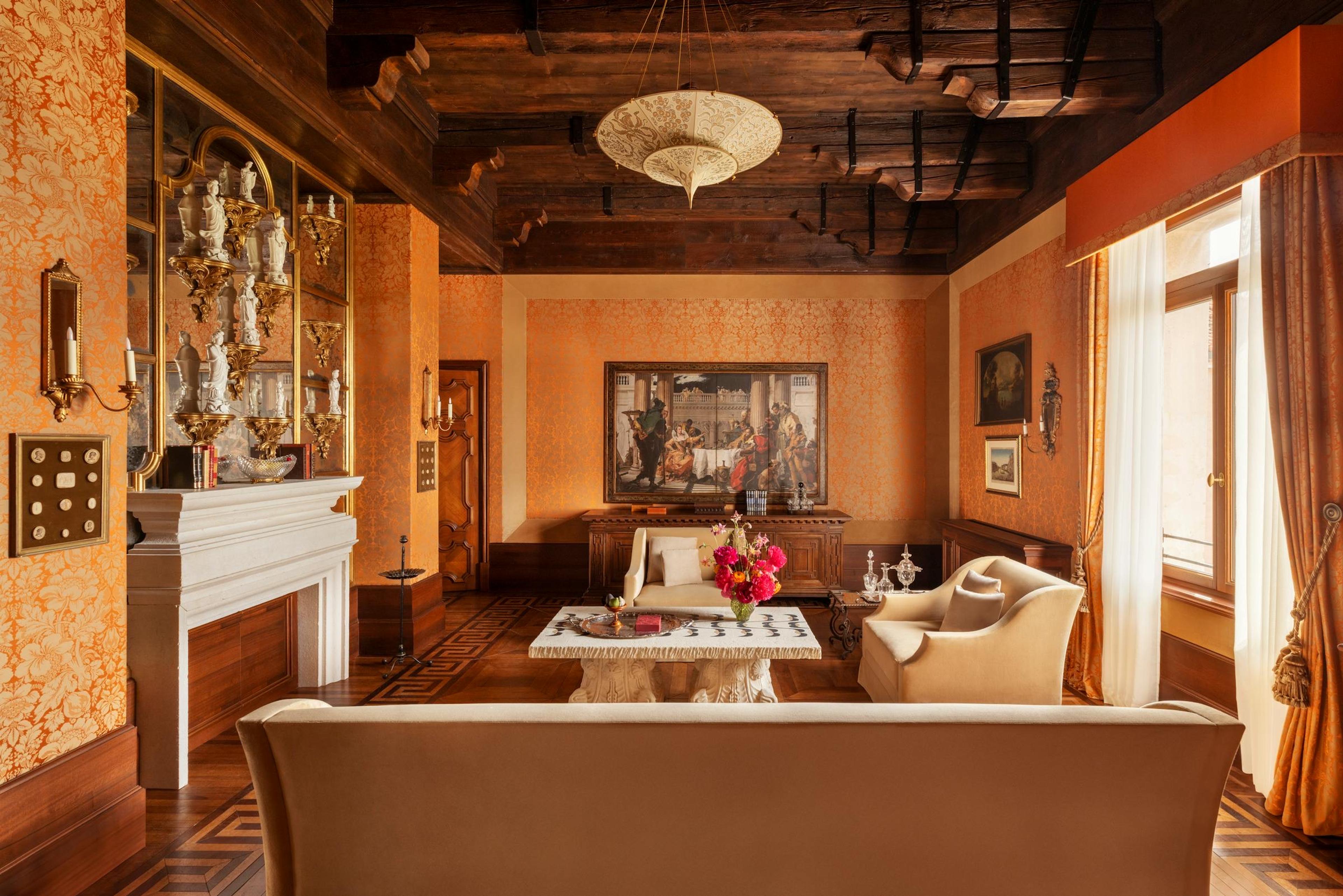 A sitting room with orange damask wallpaper, a beamed wooden ceiling, a marble fireplace, mirrored cabinets of porcelain figures, and cream armchairs around a carved stone table with red flowers.