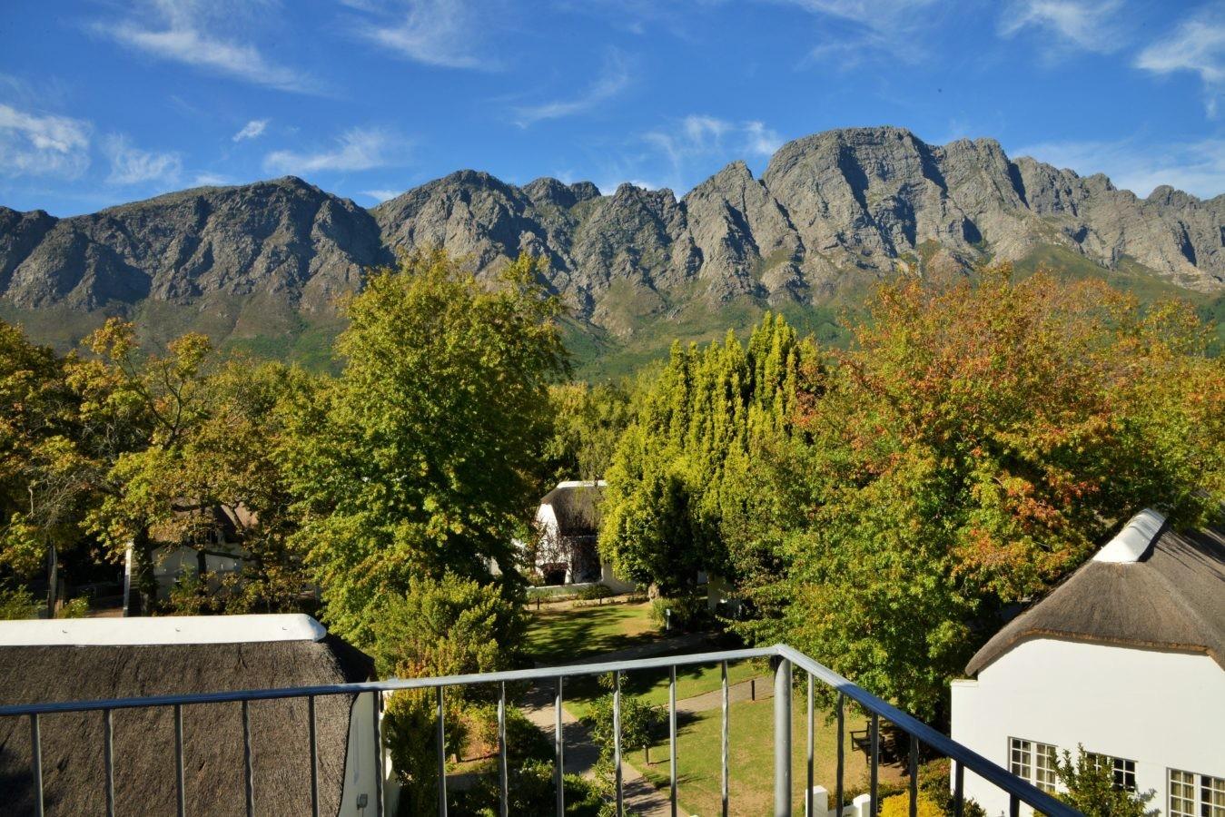 A view from a balcony over the tops of trees and thatched roofs to a long rocky mountain range under blue sky.