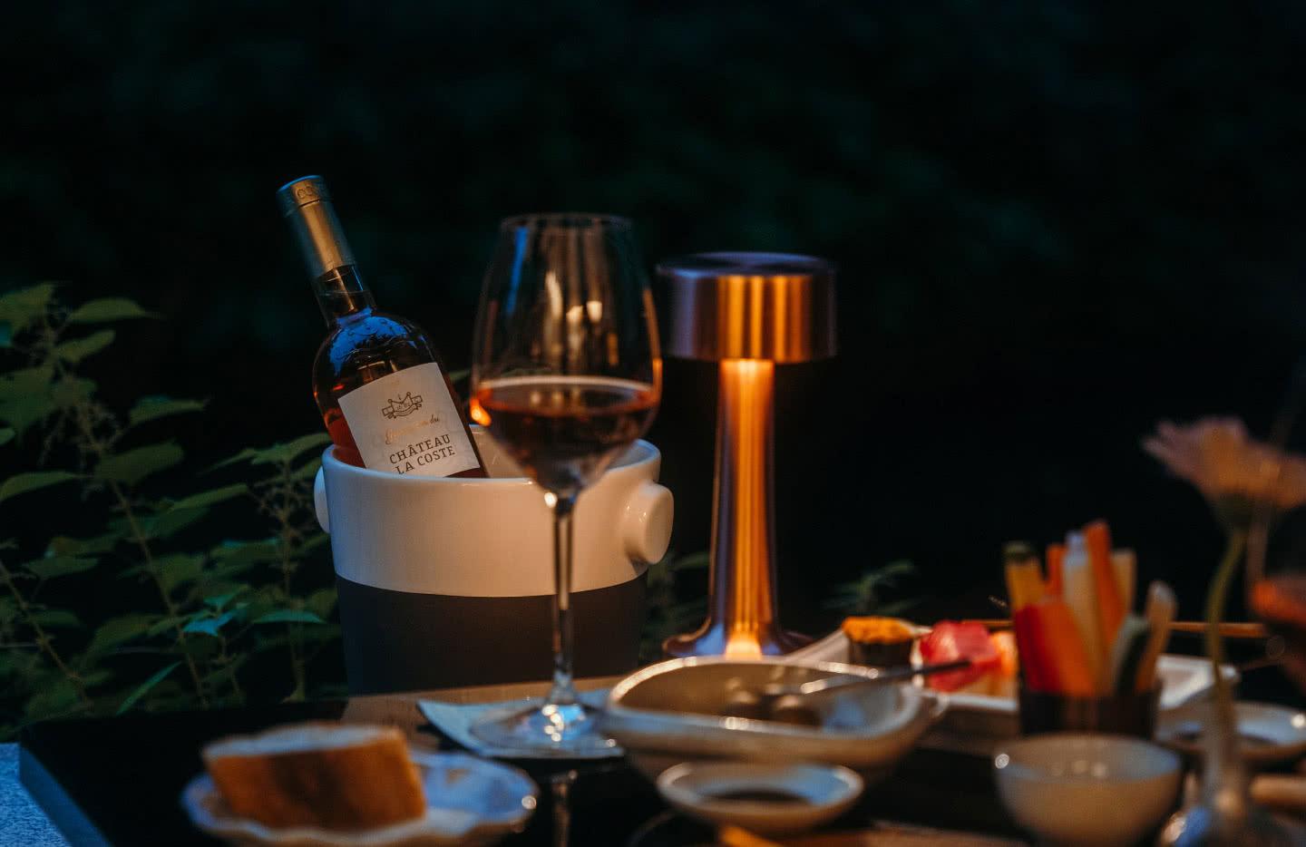 a close-up at night of a wine bottle in an ice bucket, a filled glass, and small dishes of food on an outdoor table