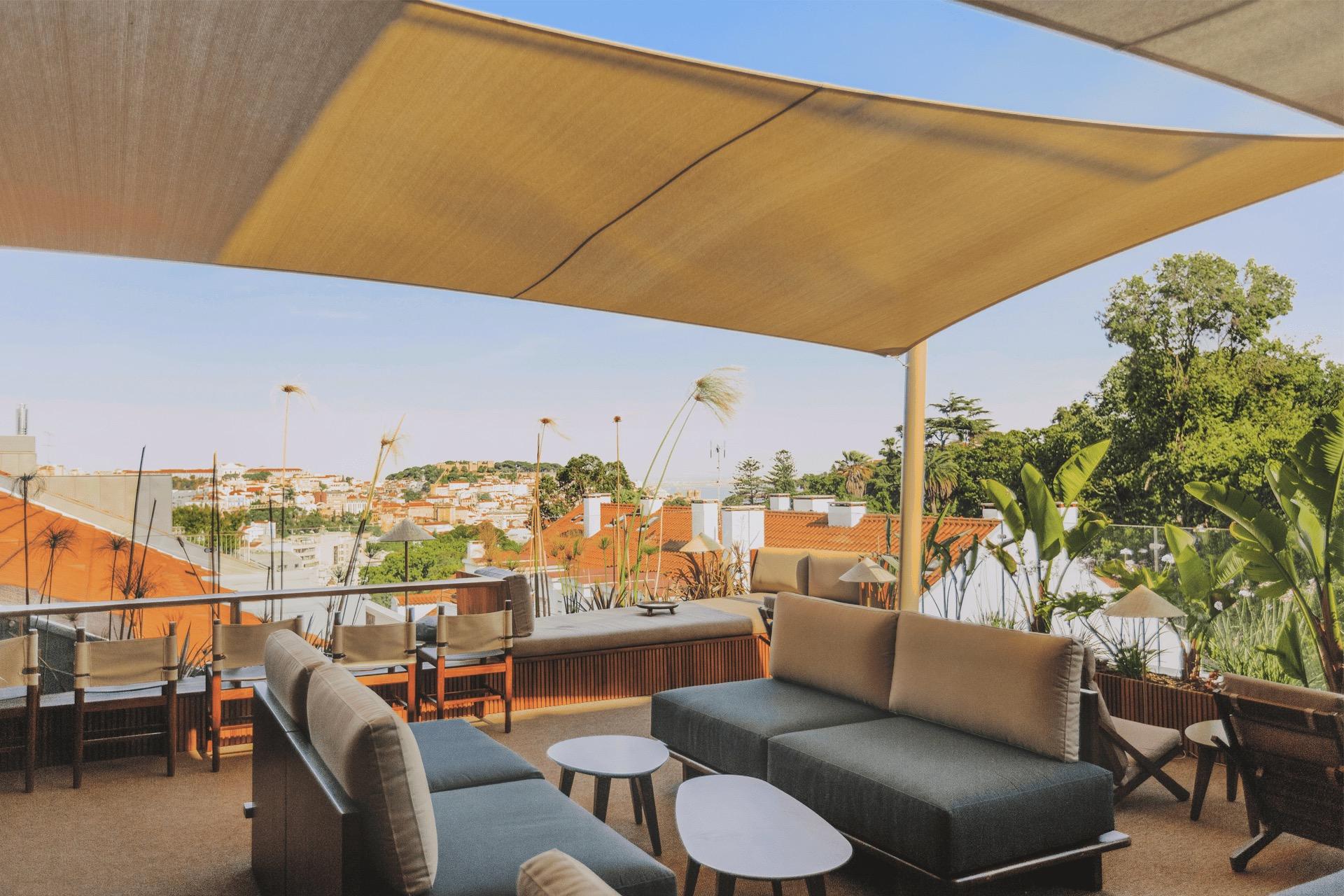 A covered terrace with grey sofas and low tables, looking over terracotta rooftops and trees towards the city.
