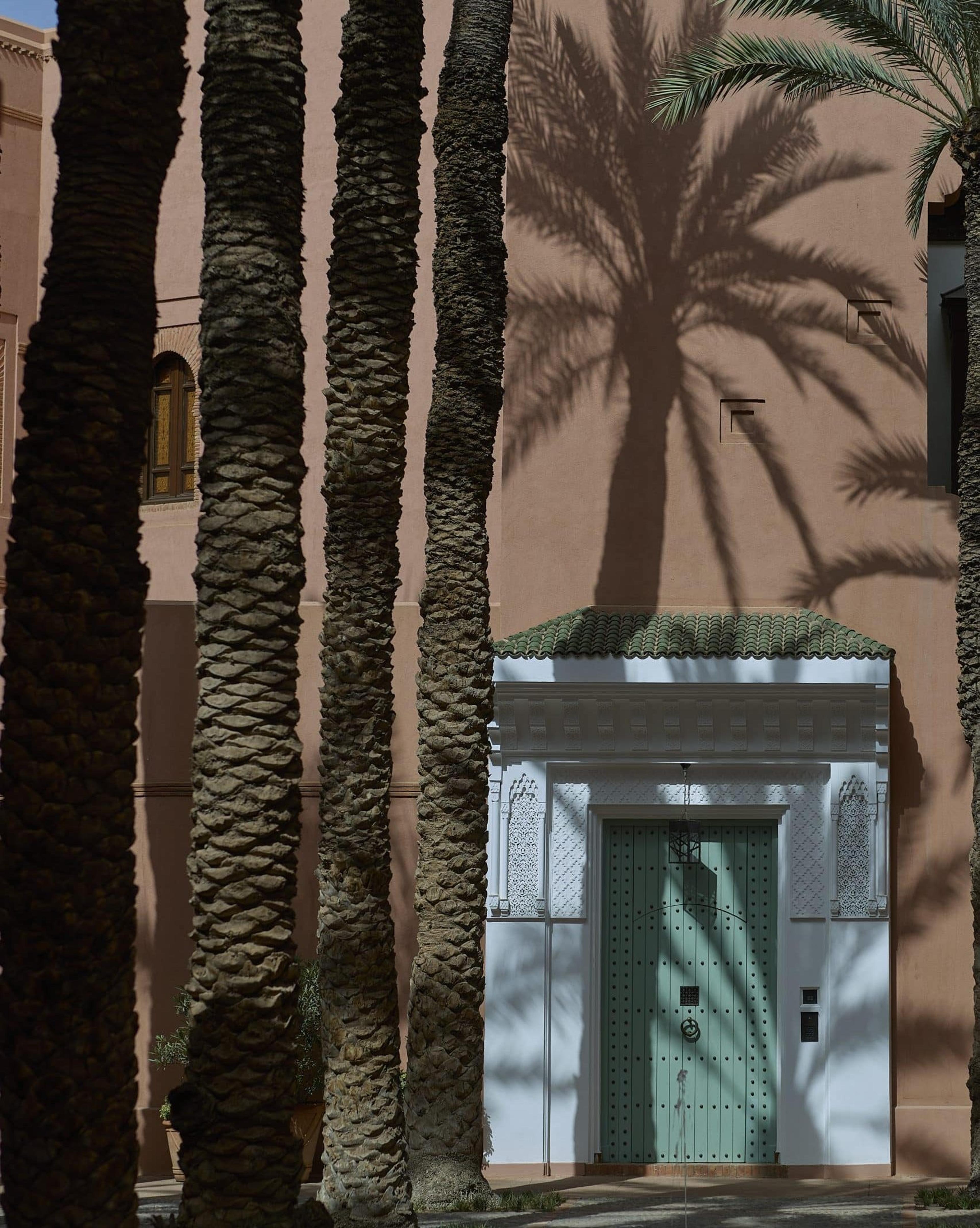 Palm trunks in front of an ochre building with a pale green door and a tiled canopy.
